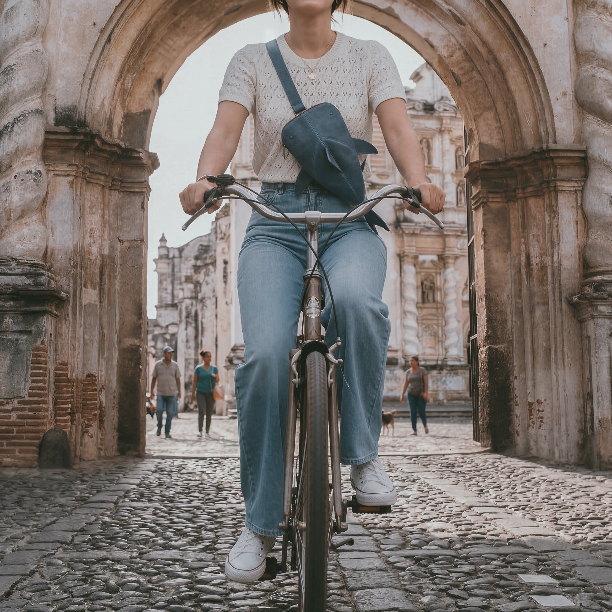 A person riding a bicycle in an urban setting, sporting the Leather Shark Crossbody Bag by Hide & Drink. This Leather Shark-Shaped Sling Bag adds a playful touch while providing comfort and functionality with its adjustable strap. Made from full grain leather, it's perfect for daily use, designed with artisan craftsmanship in La Antigua Guatemala & Oaxaca Mexico. A great choice for those wanting a stylish, practical messenger bag.