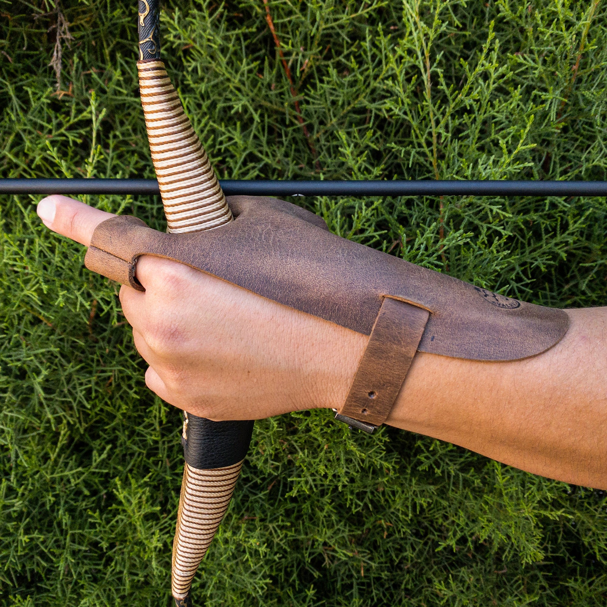An archer’s hand gripping a bow, outfitted with the Valhalla Gear Archery Shooting Glove. This glove acts as a Hand Guard and Wrist and Finger Protector, providing enhanced grip and comfort for using a Recurve Bow. Made with Full Grain Leather in a small batch production, it’s a stylish addition for shooting or cosplay.