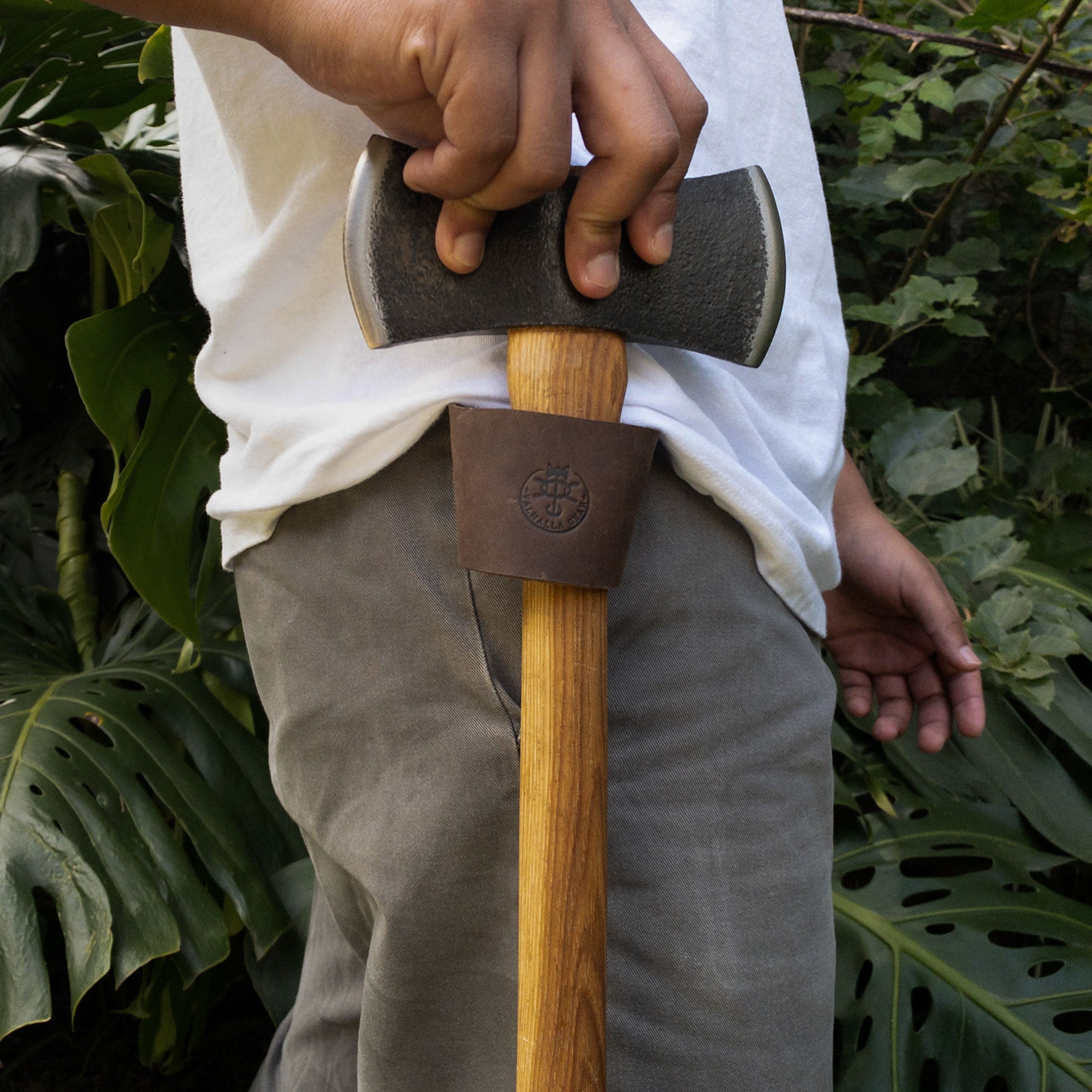 In this image, a user demonstrates Valhalla Gear's Single Axe Holster with an axe securely held at their side, highlighting the functionality and ease of this accessory for tools. Made of durable full grain leather, this axe holster is perfect for safe transport during home improvement projects, representing artisan craftsmanship from La Antigua Guatemala & Oaxaca Mexico with double belt loops for added convenience.