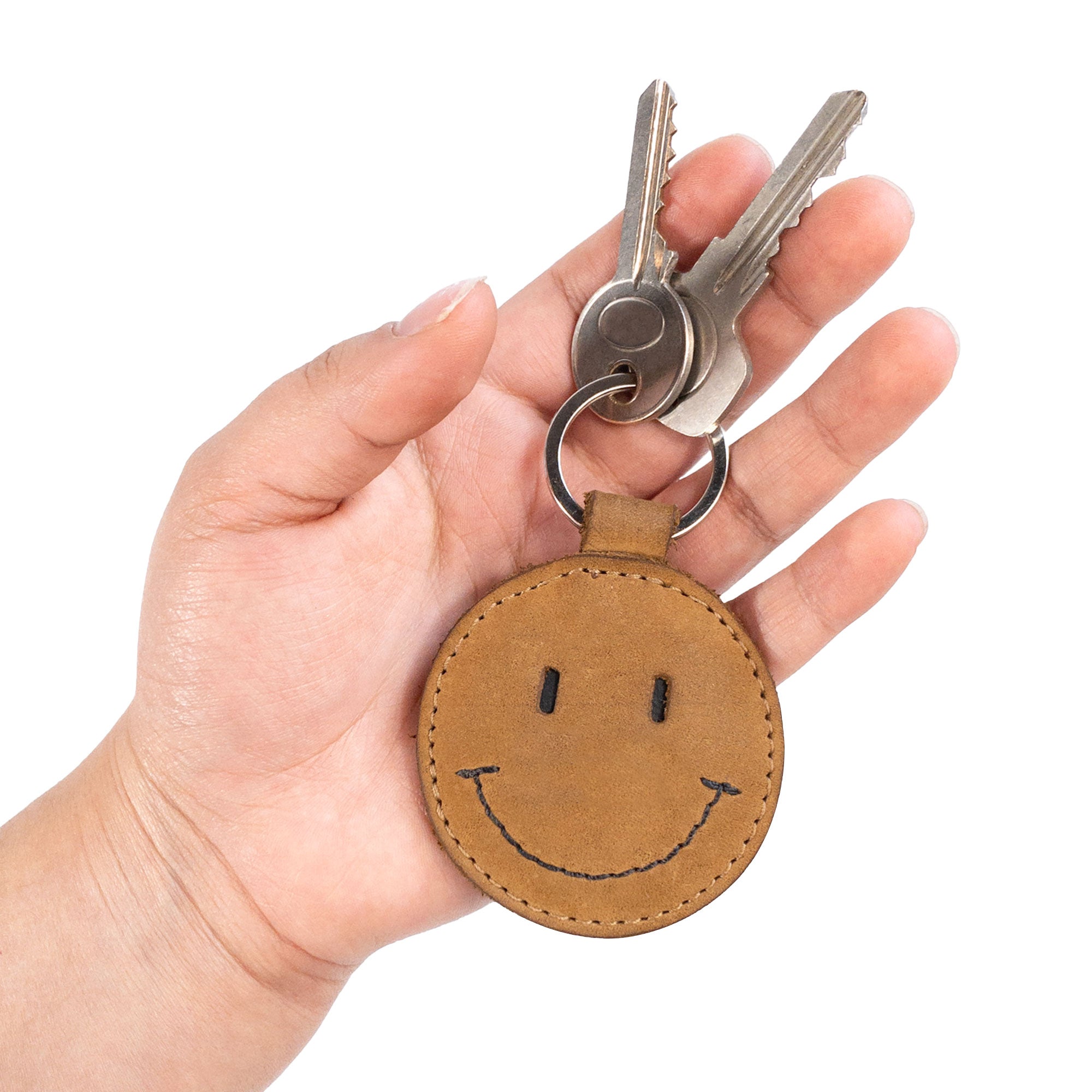 A hand holding the Smiley Face Keychain by Hide & Drink, highlighting its fun and stylish design. This key ring is made of high-quality Full Grain Leather, adding a unique touch to everyday accessories. Perfect for gifting and showcasing artisan skills from Oaxaca Mexico, it's a great example of handmade craftsmanship.