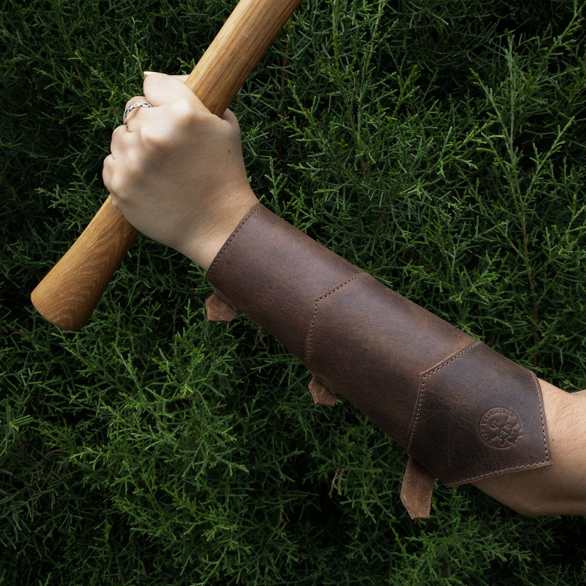 A powerful grip on a wooden staff, showing the Valhalla Gear Forearm Protector for Bow Shooting Practice worn as a Female Archery Bracer. This sturdy armband, designed with adjustable buckle straps, emphasizes the characteristics of high-quality full grain leather, perfect for cosplay and outdoor sports, along with its handmade origins from artisan craftsmen in La Antigua Guatemala.