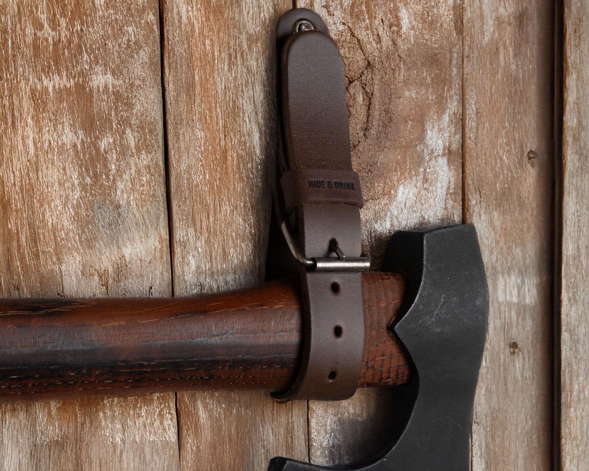 Close-up of the Hide & Drink Wall Hanging Strap securing an axe against a rustic wooden surface. This image showcases the durability and secure fit of this heavy-duty tool hanger. With a buckle closure suitable for axes, hatchets & tools, these wall hanging straps are crafted from full grain leather and are handmade with pride, emphasizing small batch production in La Antigua Guatemala & Oaxaca Mexico.