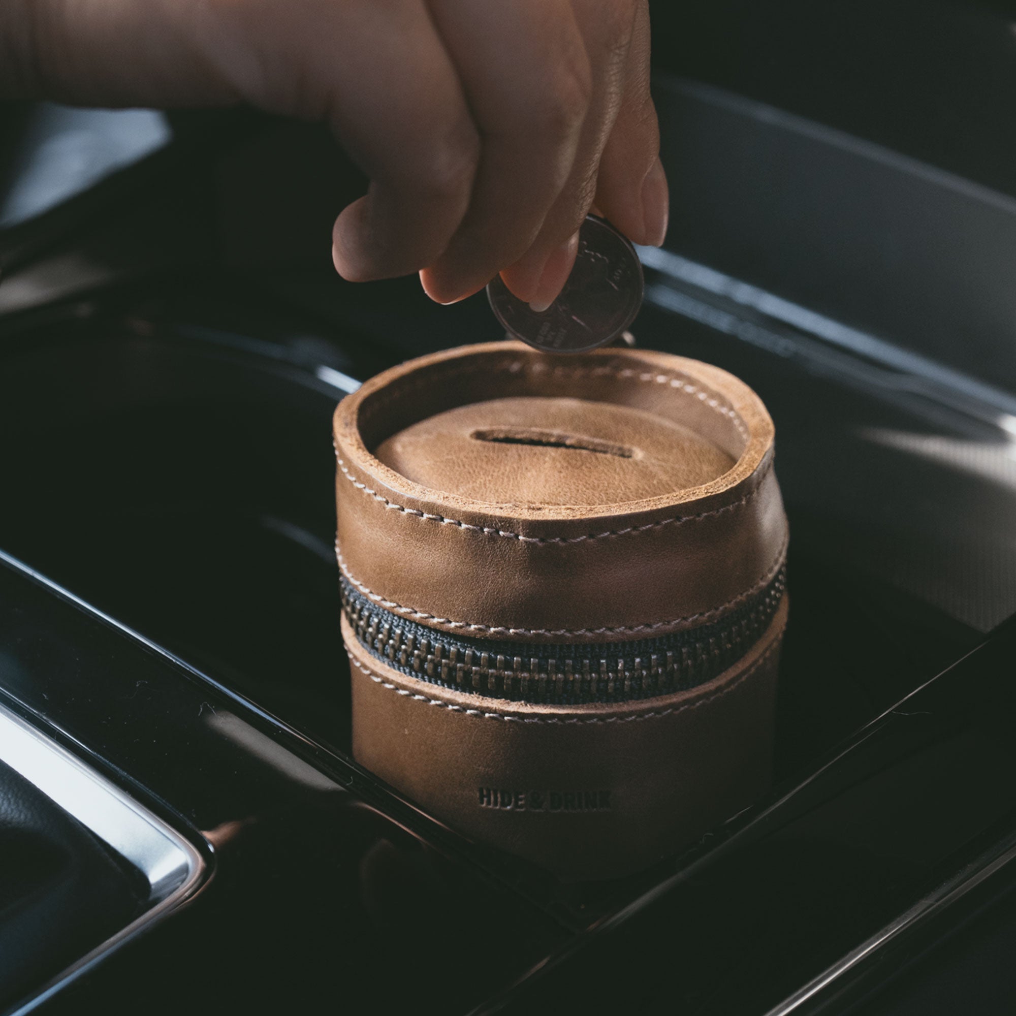 A hand placing a coin into the Bucket Coin Release by Hide & Drink, showcasing its practical use as a Money Box. This Full Grain Leather Piggy Bank Bucket fits perfectly in car cup holders, demonstrating its role as an artisan-crafted Cash Organizer, handmade in La Antigua Guatemala & Oaxaca Mexico.