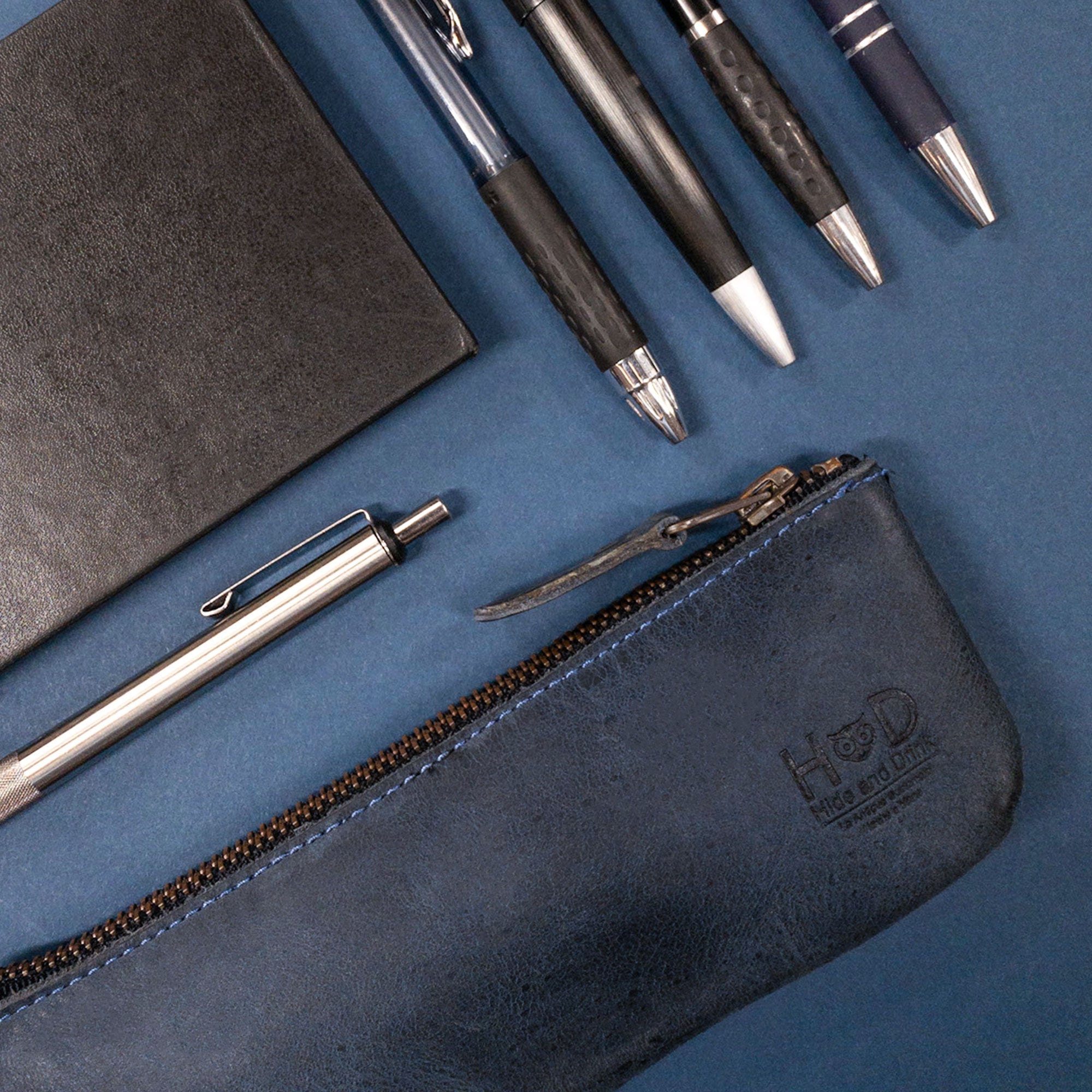 Hide & Drink Pencil Pouch displayed with pens on a blue backdrop, showcasing its elegant design and practical use as a pen and marker pouch for men and women. The full grain leather highlights the classic style for artists, lovingly handcrafted in small batches in La Antigua Guatemala & Oaxaca Mexico, making it a unique addition to your collection.