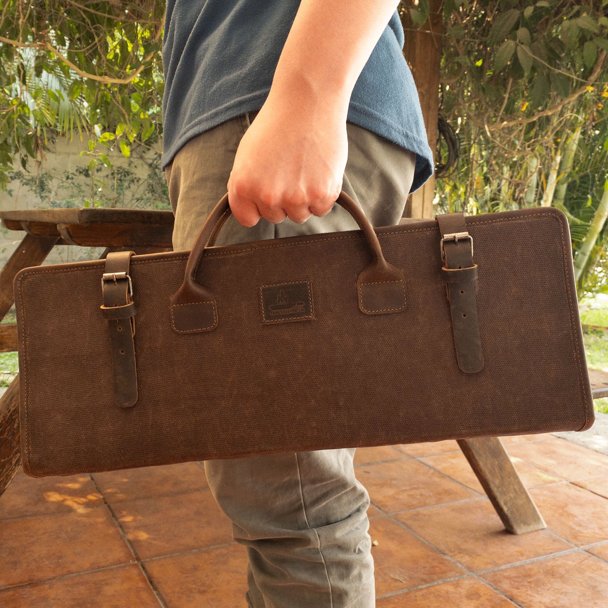 Person holding Cast Leather Co. Grill Tool Holder (Tools NOT included), a portable carry bag designed for BBQ grilling accessories. This Grill Tools Set Case combines full grain leather and waxed canvas for outdoor grill enthusiasts. Hand cut and hand sewn, this artisan piece highlights small batch production crafted in La Antigua Guatemala and Oaxaca Mexico.