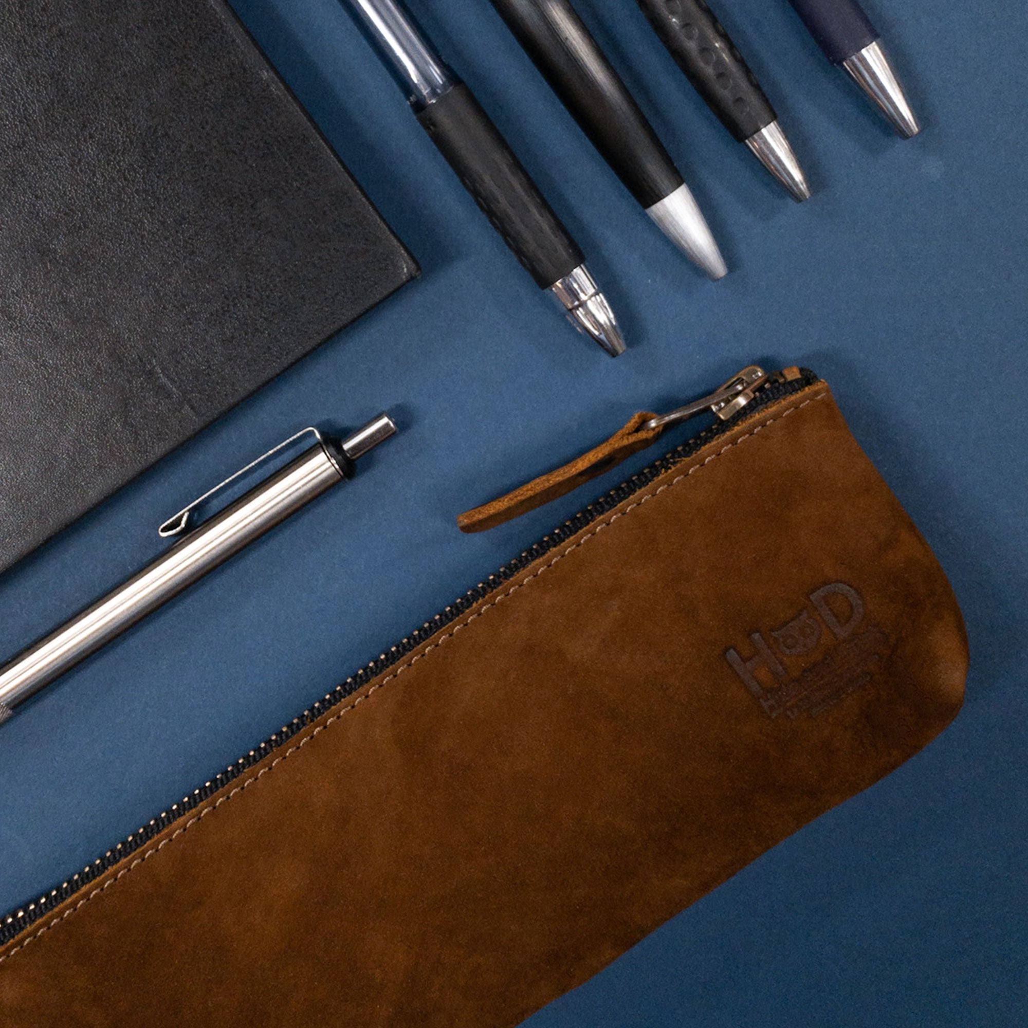 Hide & Drink Pencil Pouch with pens and a notebook, emphasizing its full grain leather construction. This rectangular pencil case functions as a stylish pen & marker pouch for men and women, featuring a flat zippered holder. Handmade in La Antigua Guatemala & Oaxaca Mexico, known for artisan craftsmanship.