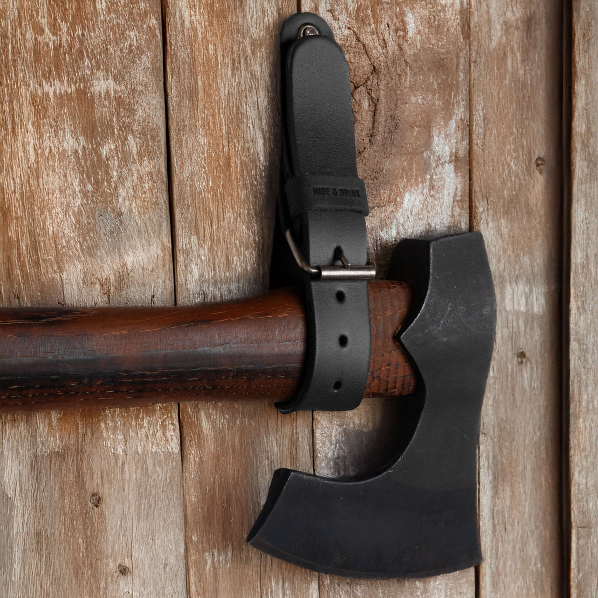 Close-up image of a black Hide & Drink Wall Hanging Strap holding an axe against wood, highlighting its sturdy and reliable craftsmanship. Featuring a buckle closure, this heavy-duty tool hanger adds style and organization, making it great for axes, hatchets & tools. Made from full grain leather, it is handmade, showcasing artisan quality from La Antigua Guatemala & Oaxaca Mexico.