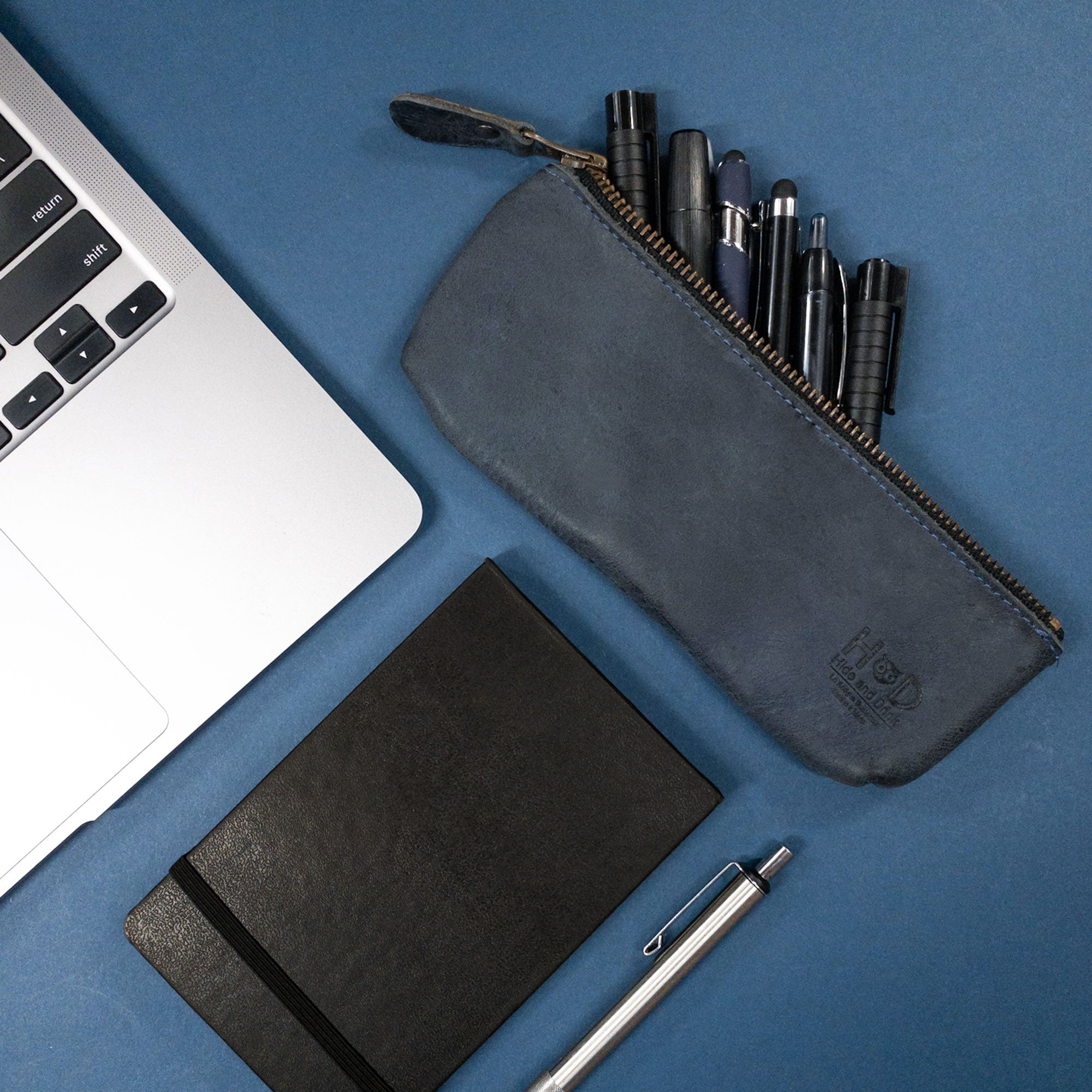 Hide & Drink Pencil Pouch beside a notebook and laptop, illustrating its versatility as a flat zippered holder perfect for storing pens and markers. The beautiful full grain leather represents classic style for both men and women, and each pouch is artisan-made in La Antigua Guatemala & Oaxaca Mexico for a touch of unique craftsmanship.