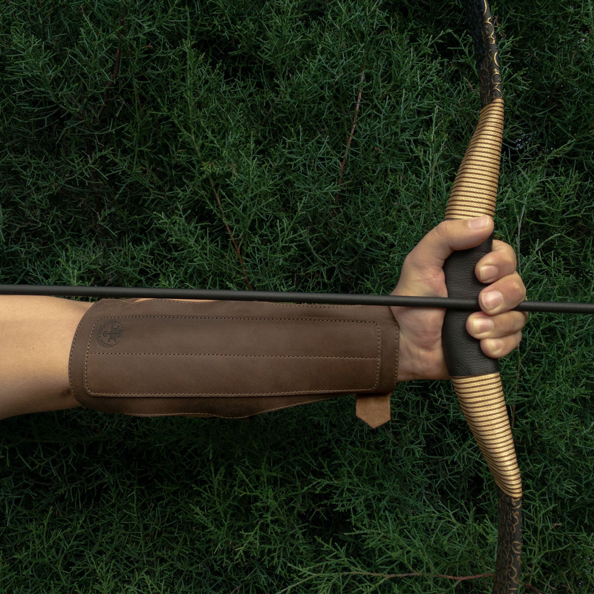 A hand gripping a bow with the 3-Buckle Archery Bracer from Valhalla Gear protecting the forearm. This handmade leather bracer is designed for archery enthusiasts, providing comfort and style during shooting practice. Perfect for those seeking quality craftsmanship.