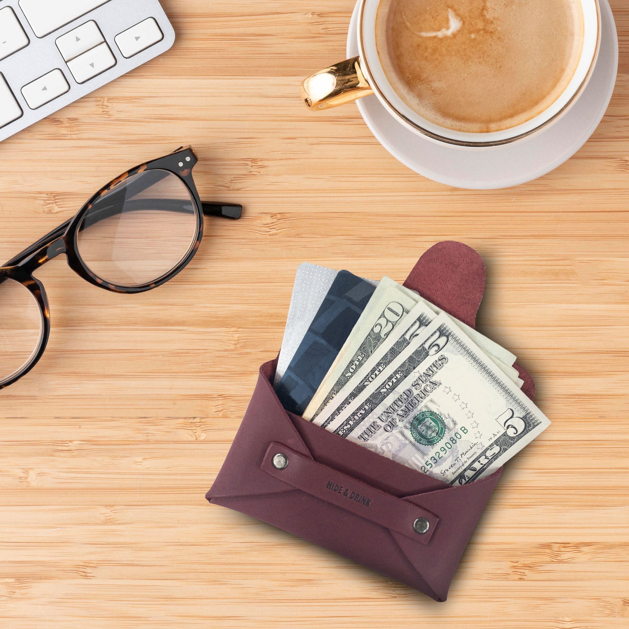 Hide & Drink's Snapless Business Card Holder on a desk with coffee. This chic wallet serves as a versatile Snapless Business Card Holder for Men and Women, providing a sturdy pouch and money organizer. Expertly made of Full Grain Leather, this product showcases small batch production with hand cut, hand sewn, and fire branded details from artisans in La Antigua Guatemala & Oaxaca Mexico.