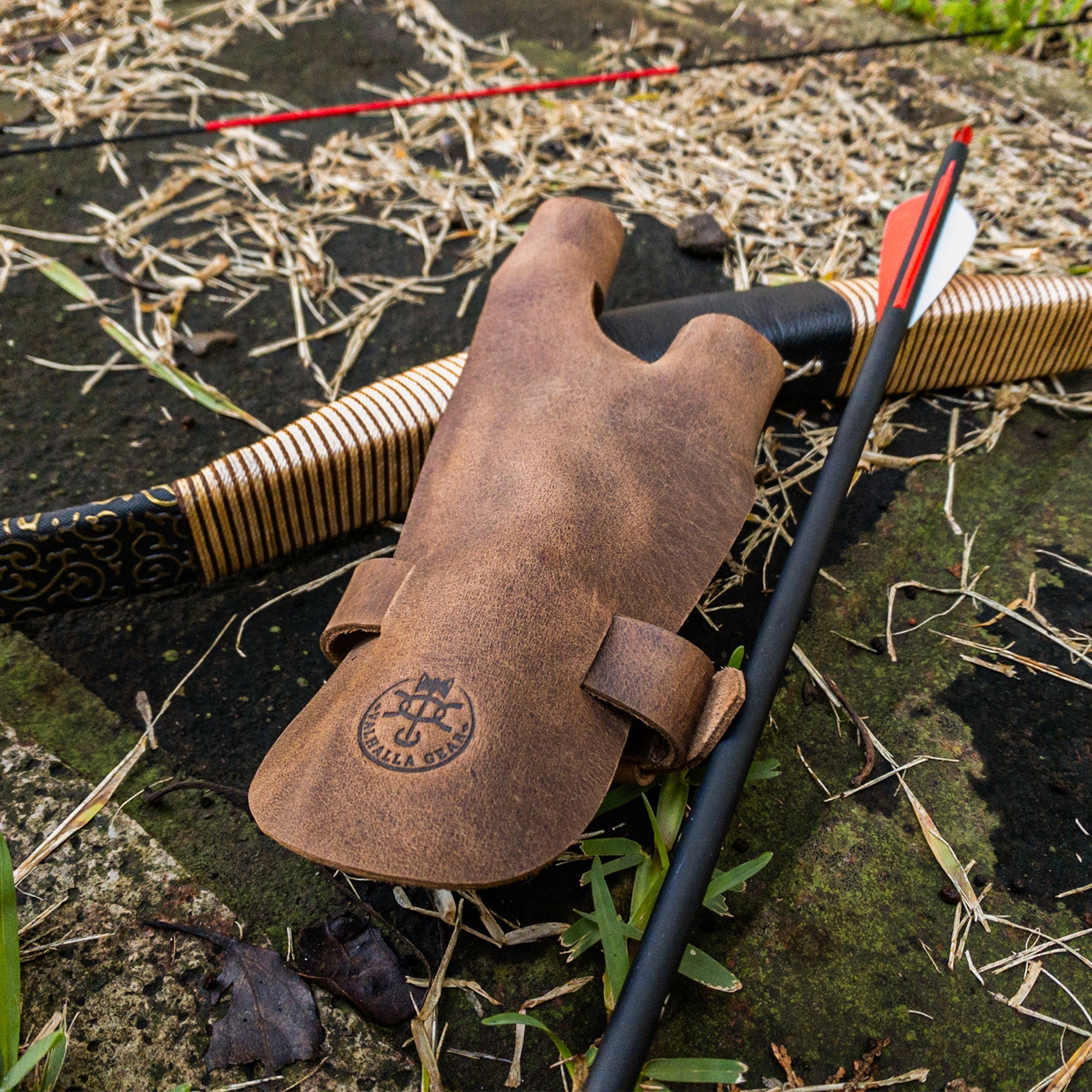 The Valhalla Gear Archery Shooting Glove placed on grass alongside an arrow and bow. This glove, made from Full Grain Leather, serves as a Hand Guard and Wrist and Finger Protector for archery enthusiasts using a Recurve Bow. Ideal for shooting or cosplay, this product reflects the artisan craftsmanship from La Antigua Guatemala.