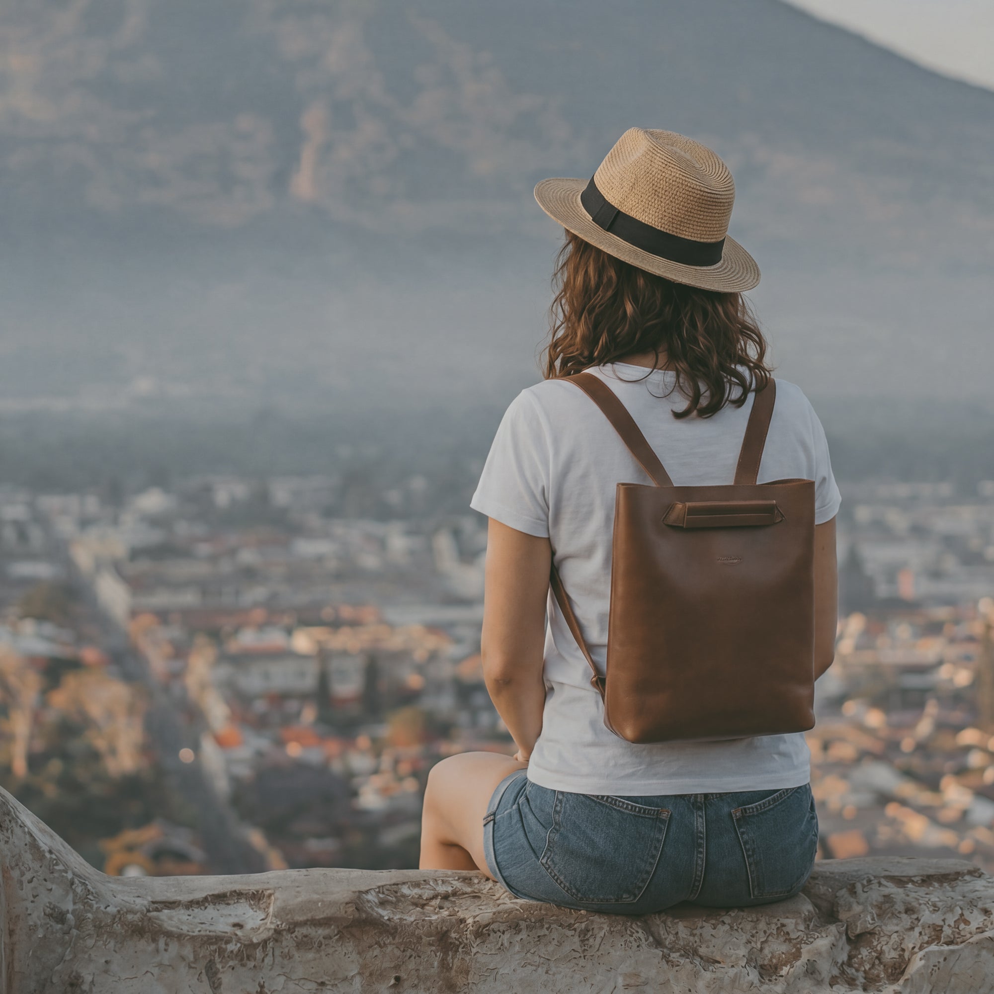 Outdoor lifestyle shot featuring a woman wearing Heather's Convertible Backpack to Shoulder Bag while enjoying a scenic view. This design serves as both a backpack and a Spacious Crossbody for Women, showcasing the Squared Purse structure and the luxurious Full Grain Leather, reflecting Small Batch Production principles from hand cut and hand sewn artisans in La Antigua Guatemala & Oaxaca Mexico.