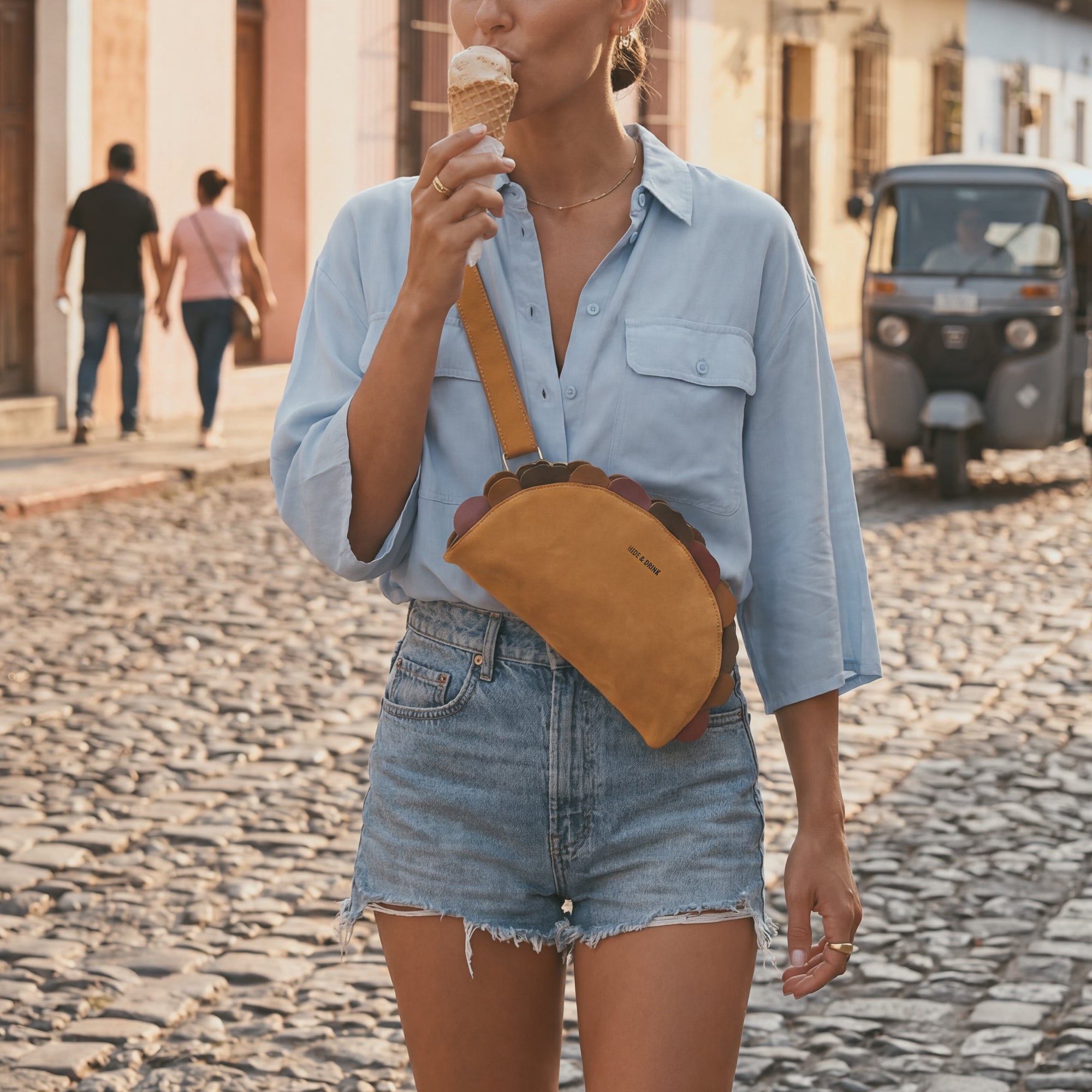 Lifestyle image of a woman enjoying ice cream while sporting the Taco-Shaped Crossbody Bag by Hide & Drink in a vibrant street setting. This Leather Taco-Shaped Crossbody Bag with Adjustable Shoulder Strap is the perfect accessory for casual outings, exemplifying exquisite craftsmanship as a messenger bag or shoulder bag, handmade in La Antigua Guatemala & Oaxaca Mexico.