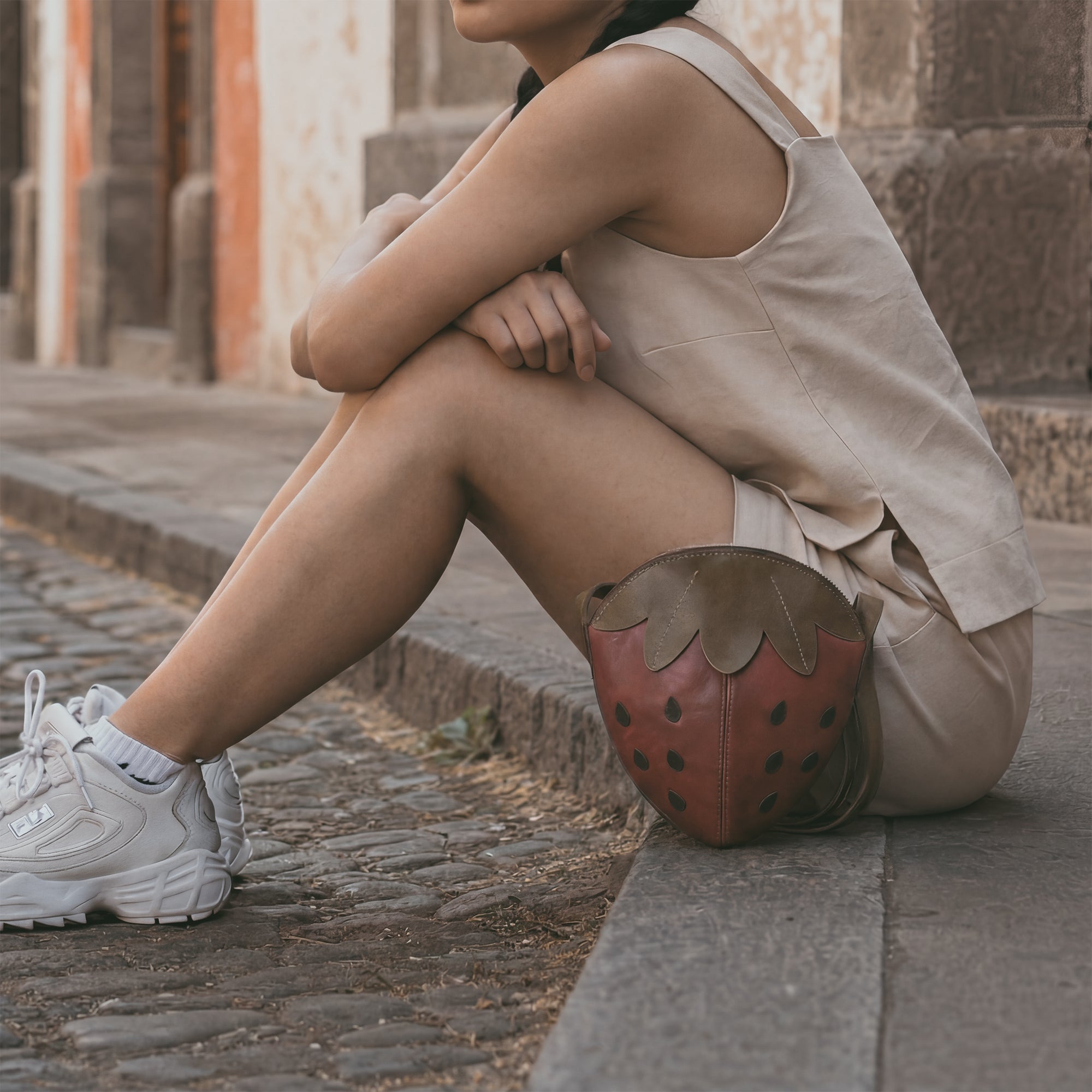 A model carrying the Leather Strawberry-Shaped Shoulder Bag by Tudi Frudi, demonstrating its stylish versatility. Made from Full Grain Leather, this crossbody bag is a must-have accessory, reflecting the quality of artisan craftsmanship from La Antigua Guatemala & Oaxaca Mexico, ensuring unique small batch production.