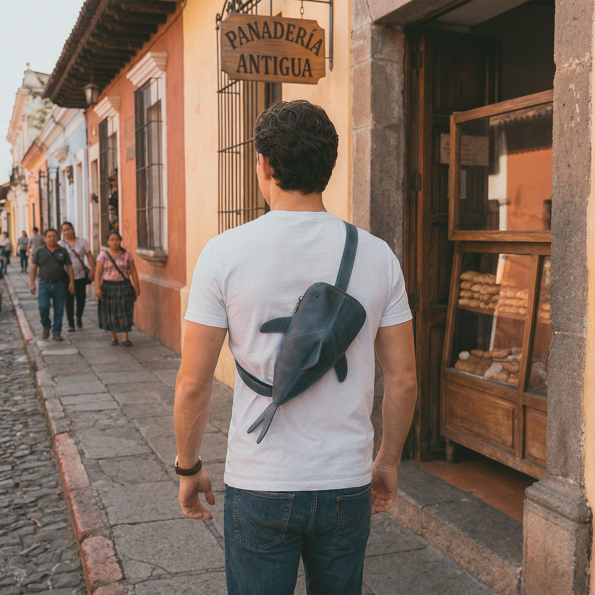 A man wearing the Leather Shark Crossbody Bag by Hide & Drink, walking through a vibrant street. The Leather Shark-Shaped Sling Bag adds fun and functionality to any outfit, designed with an adjustable strap for comfort. This artisan-crafted bag from full grain leather is perfect for day trips and casual outings, showcasing the unique craftsmanship of La Antigua Guatemala & Oaxaca Mexico.