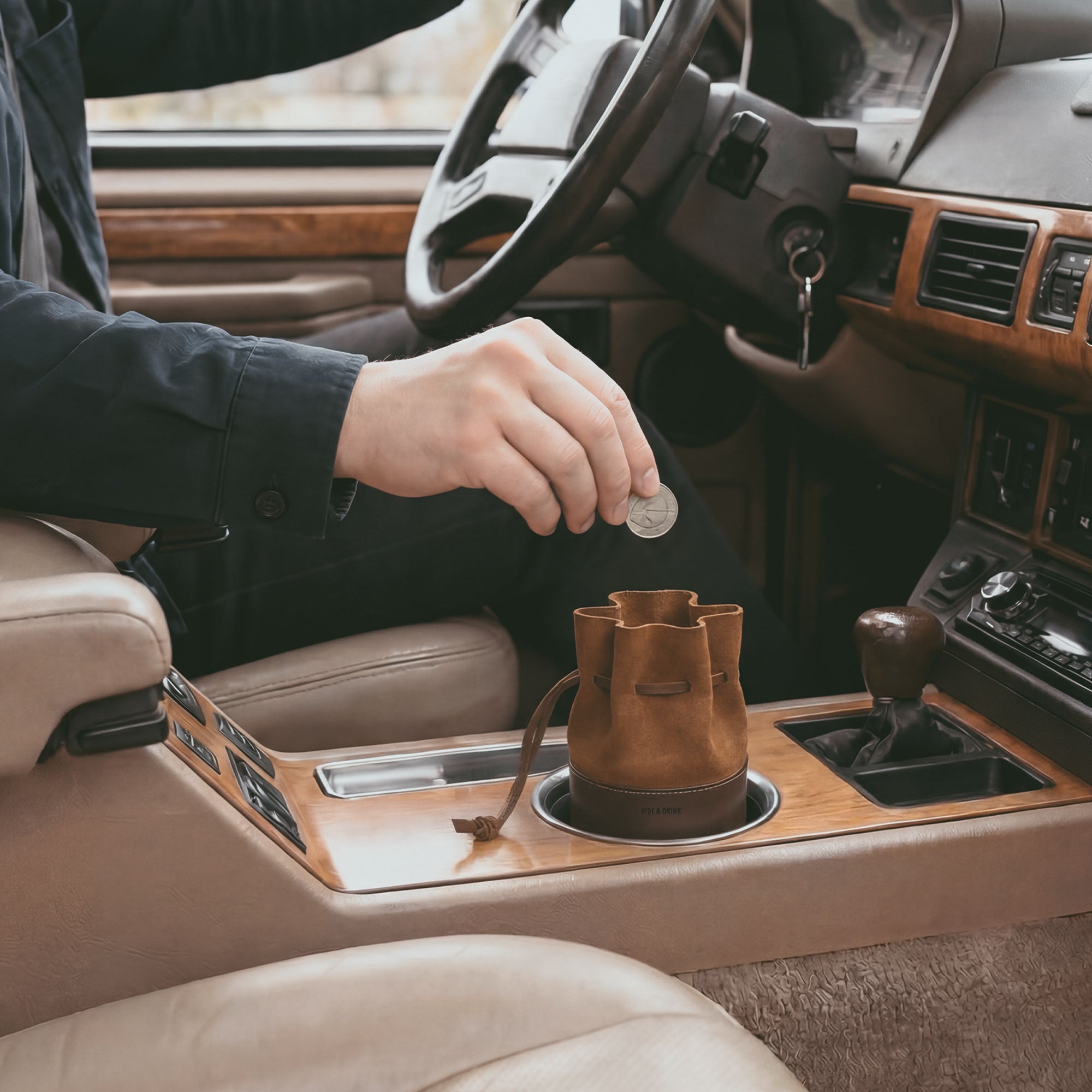 The Medieval Bucket Coin Release by Hide & Drink being used in a car showcases its functionality as a convenient piggy bank bucket. This coin pouch serves as a practical cash organizer and cylinder case for pennies, expertly hand cut and sewn from full grain leather in La Antigua Guatemala & Oaxaca Mexico.