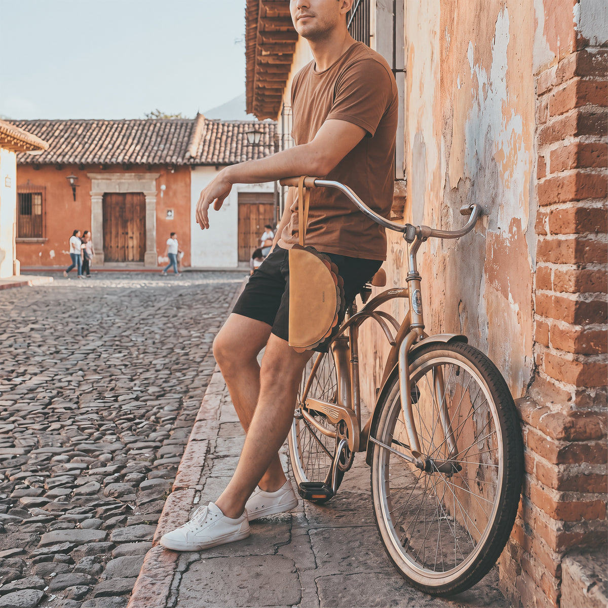 Man wearing the Hide & Drink Taco Clutch Bag while standing beside a bicycle on a cobblestone street; Taco Clutch Bag Full Grain Leather by StockyardX, featuring Small Batch Production, Hand Cut, Hand Sewn, Fire Branded Artisan craftsmanship, Handmade in La Antigua Guatemala & Oaxaca Mexico.
