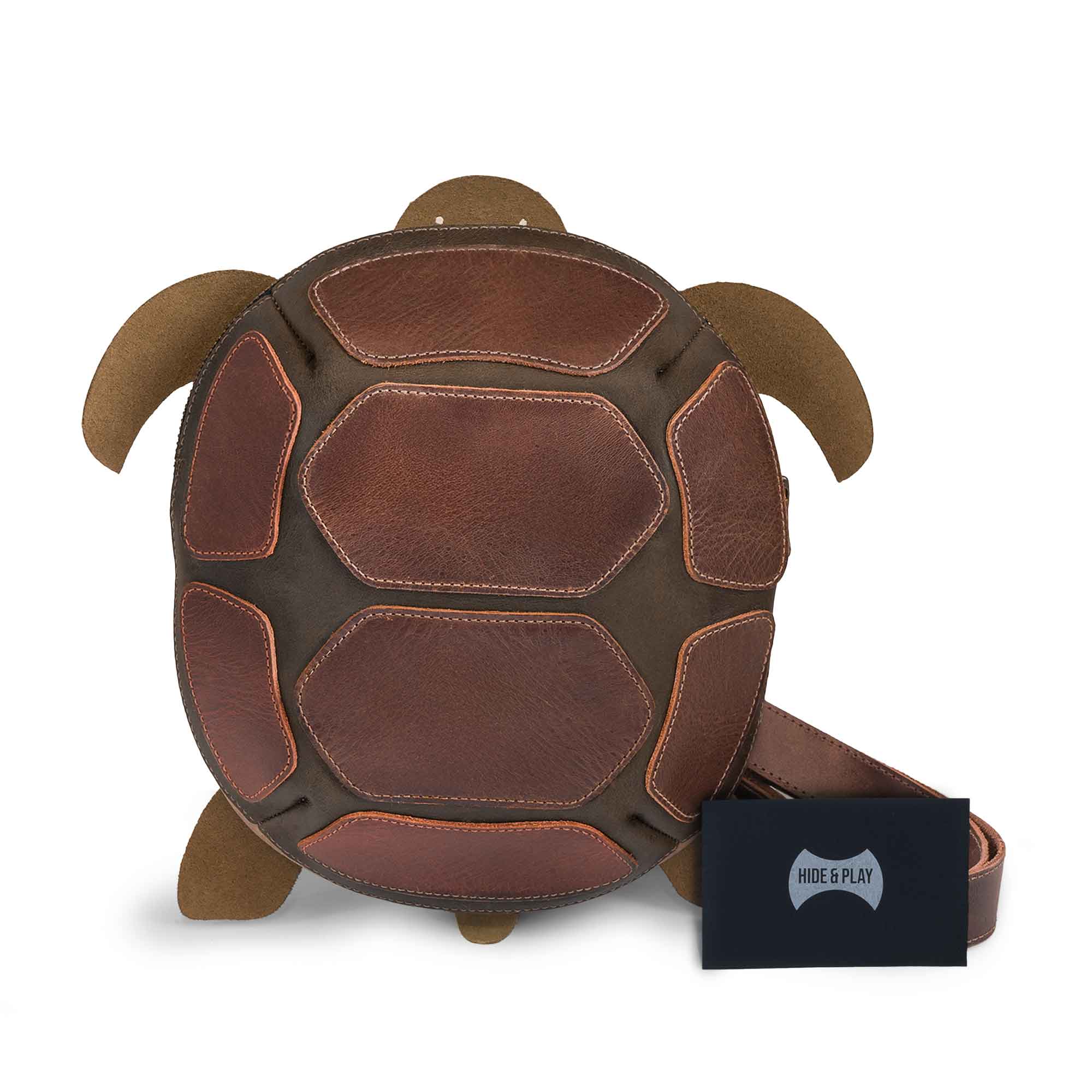Front view of the Leather Turtle-Shaped Crossbody Bag by Hide & Play alongside an authentic branded tag. This turtle design bag serves as a versatile messenger and shoulder bag, made from premium full grain leather. Handcrafted in La Antigua Guatemala & Oaxaca Mexico, it reflects small batch production and artisan quality.