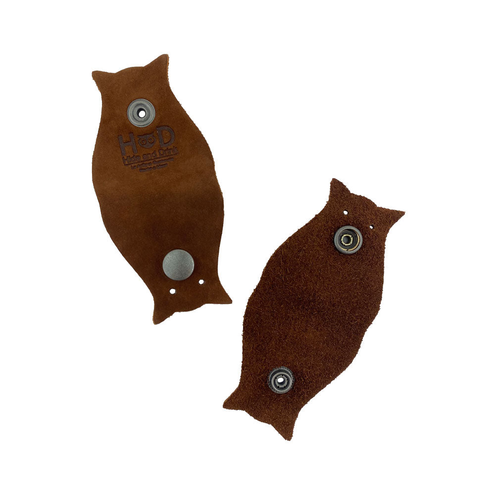 Owl Shaped Cord Keeper (2-Pack) by Hide & Drink emphasizing the leather's quality with the brand's logo on each piece. Owl Shaped Cord Keeper (Cord Clam) 2-Pack. Full Grain Leather, StockyardX, Small Batch Production, Hand Cut, Hand Sewn, Fire Branded, Artisan, Handmade in La Antigua Guatemala & Oaxaca Mexico.