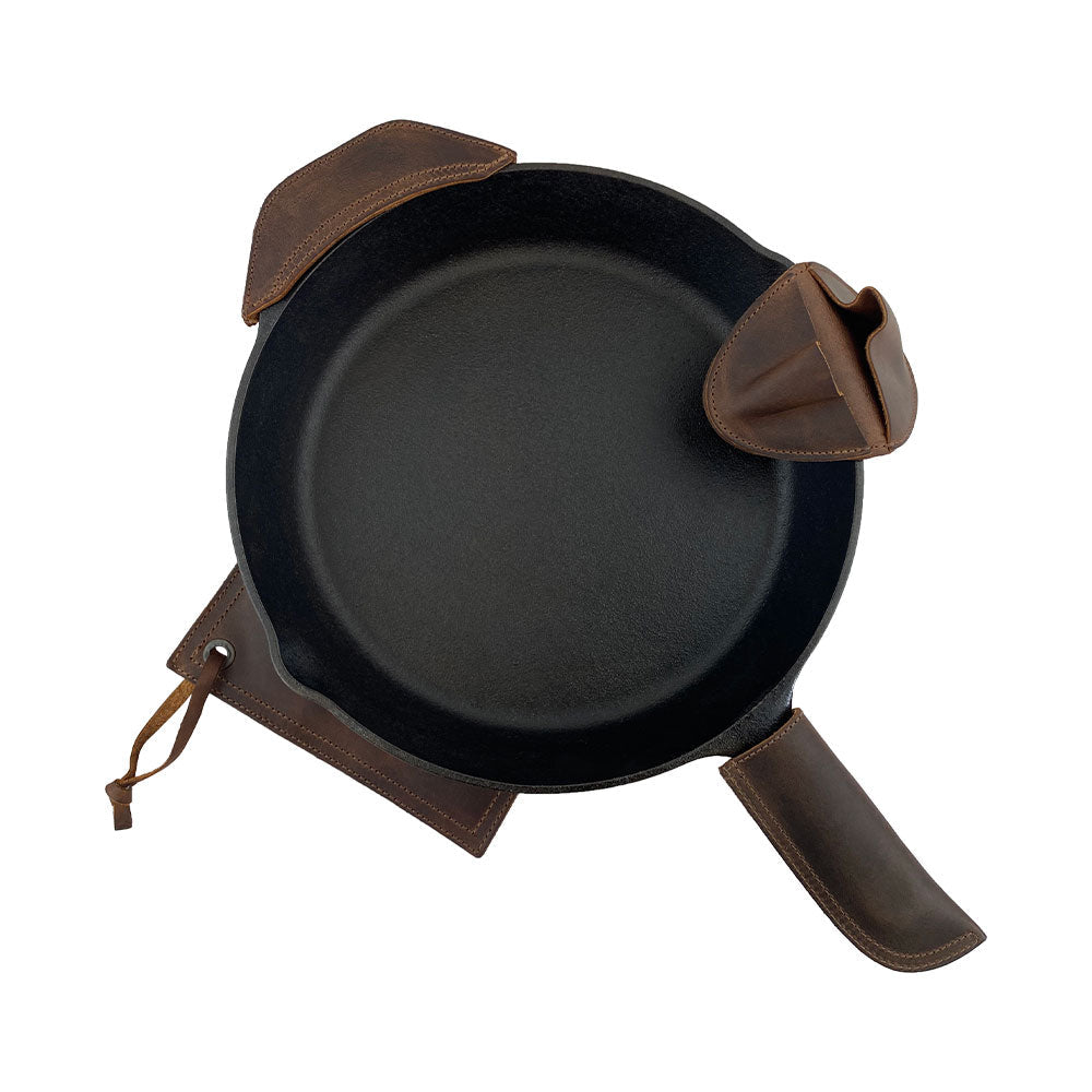 The Hide & Drink Hot Handle Holders (Set of 4) are presented with a cast iron skillet, featuring the Panhandle and Side Kick alongside the Grip Mitt and Square Pot Holder. Constructed from premium Full Grain Leather, these handmade items are crafted in small batch production, showcasing hand cut, hand sewn quality by artisans in La Antigua Guatemala & Oaxaca Mexico.