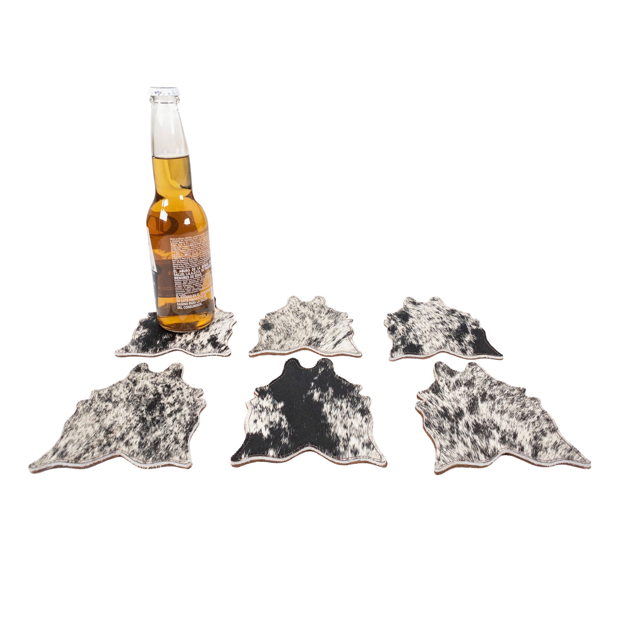 Stylish black and white Cowhide Shape Set of 6 Coasters from Western Leather Works displayed with a beer bottle. These handcrafted coasters, made from Hair On Hide Full Grain Leather, are ideal for drinks and embody the artisan spirit of La Antigua Guatemala & Oaxaca Mexico.