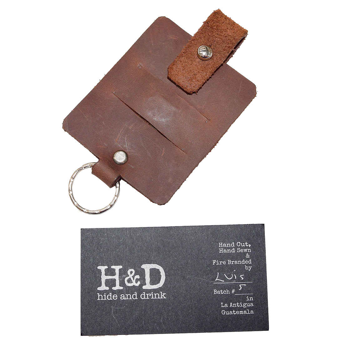 Hide & Drink Cable Holder with Key Ring accompanied by a business card featuring branding details, crafted from Thick Full Grain Leather. This unique keychain and cord organizer is perfect for anyone needing to keep their cables neat and accessible. Celebrate artisan craftsmanship with this handmade product from La Antigua Guatemala & Oaxaca Mexico.
