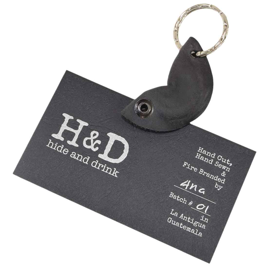 The Fidget Cashew Keychain by Hide & Drink is displayed with a business card, emphasizing its handmade quality. This key holder, designed as a Good Luck Charm, is crafted from Full Grain Leather, showcasing the artisan skills of small batch production through hand cut, hand sewn, and fire branded techniques in La Antigua Guatemala and Oaxaca Mexico.