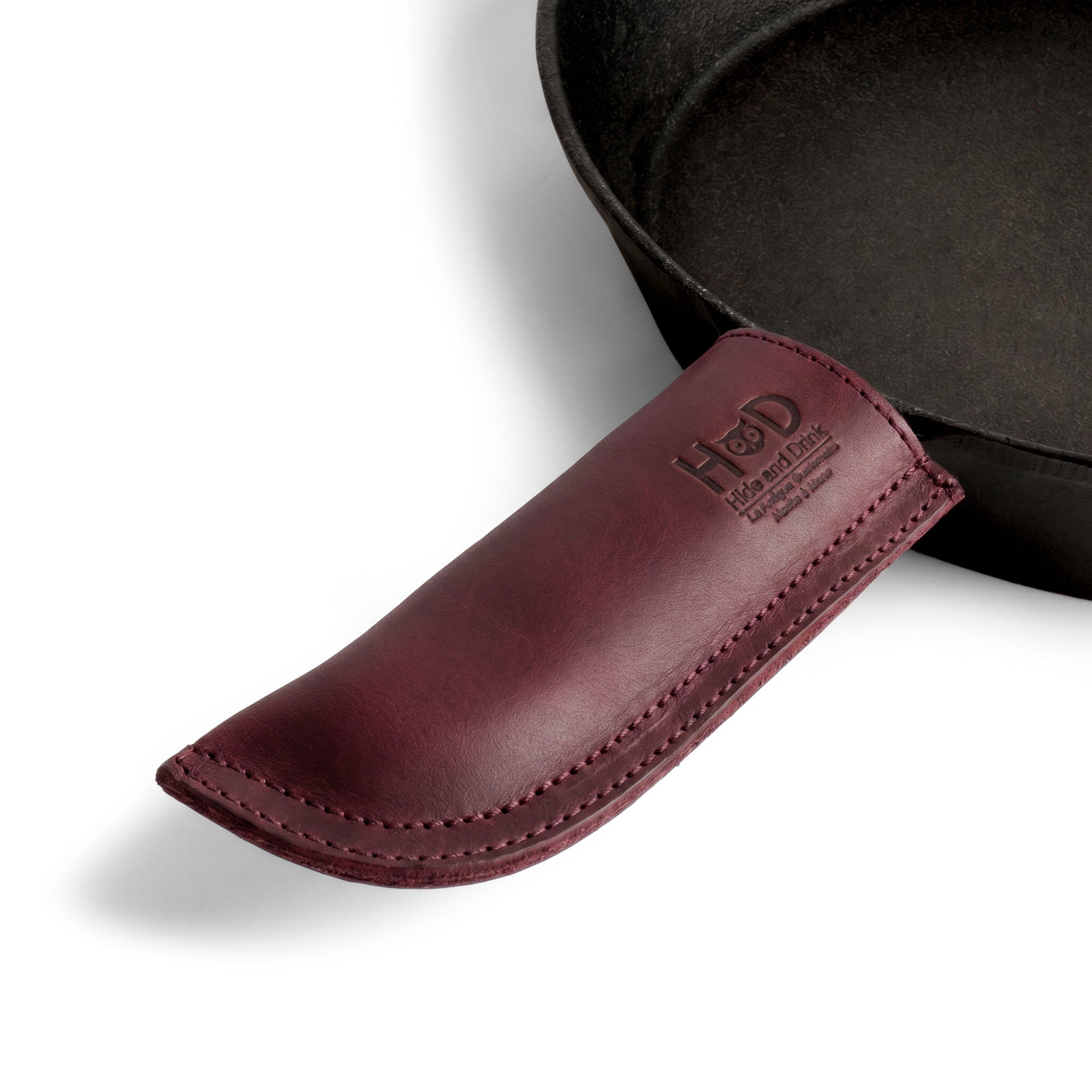 Hide & Drink Cast Iron Panhandle Cover elegantly presented in a stylish showcase. This artisan hot handle protector, crafted from durable full grain leather, features a double stitched design for added strength. Handmade in La Antigua Guatemala & Oaxaca Mexico, it's an essential tool for any kitchen.