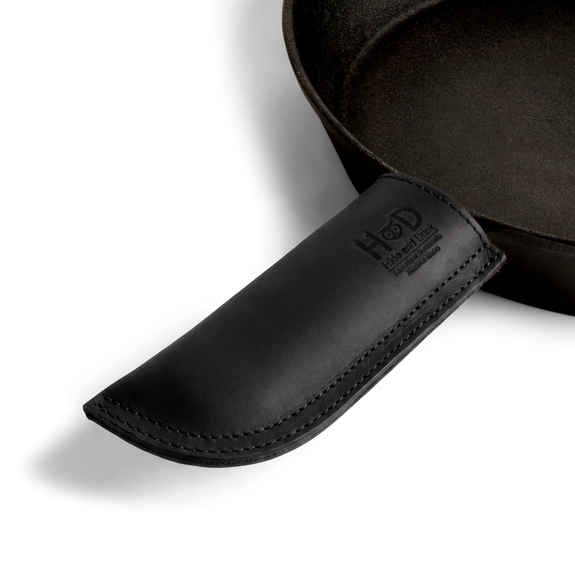 Hide & Drink Cast Iron Panhandle Cover displayed alongside its packaging, emphasizing the details. This artisan hot handle protector is made from double stitched full grain leather and provides superior heat protection. Handmade in La Antigua Guatemala & Oaxaca Mexico, it's perfect for any kitchen setup.