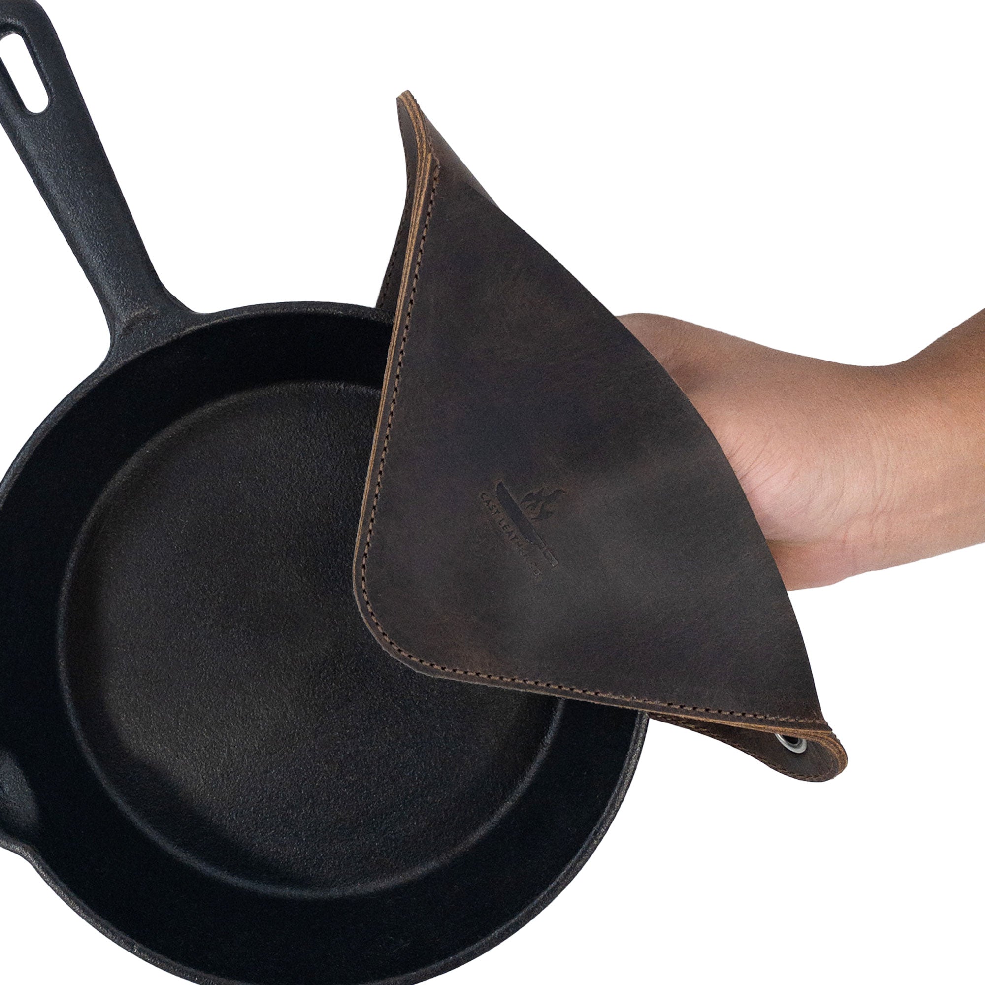 A Cast Leather Co. Leather Oven Mitt & Trivet used to grasp a hot cast iron skillet, emphasizing its fire-branded finish and robust construction. This heat-resistant accessory is designed as a squared hot pot pad (potholder) and showcases the quality of full grain leather from small batch production and artisan methods in La Antigua Guatemala.