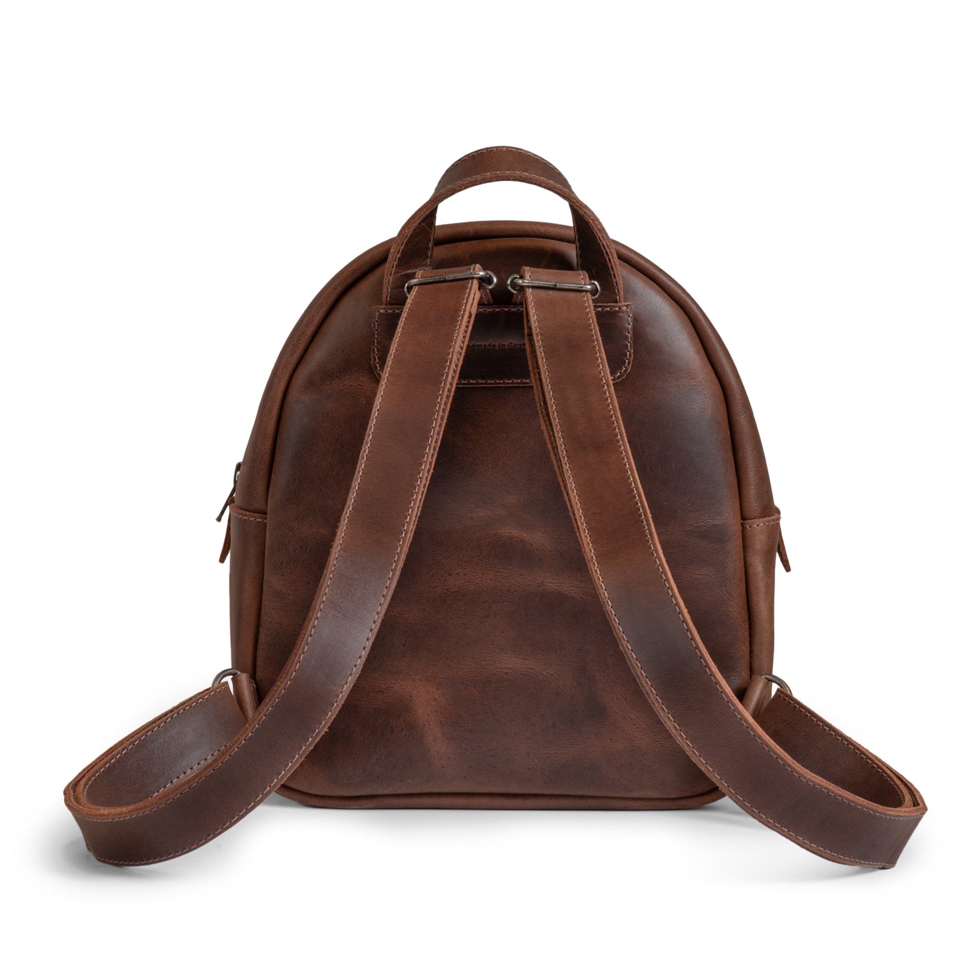 The back of The Holly Mini Backpack from Austin Genuine, highlighting its sturdy straps and exquisite full grain leather, accompanied by tags such as Full Grain Leather, StockyardX, Small Batch Production, Hand Cut, Hand Sewn, Fire Branded, Artisan, Handmade in La Antigua Guatemala & Oaxaca Mexico.