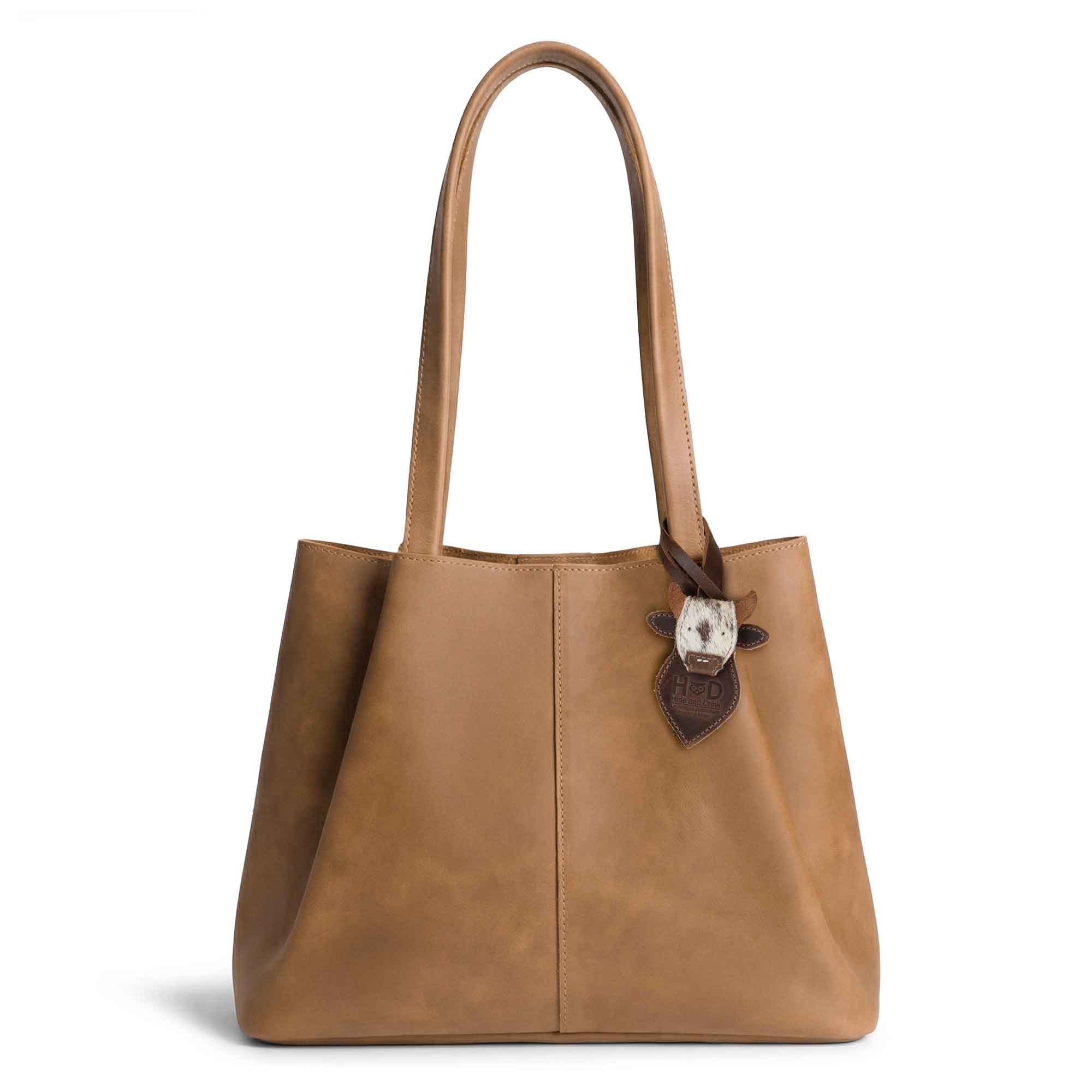 Hide & Drink Bag Charms attached to a chic brown handbag, featuring a cow head design perfect for embellishment. This purse charm for tote & backpacks made of full grain leather symbolizes StockyardX's commitment to small batch production. Each piece reflects hand cut, hand sewn techniques and is fire branded, embodying artisan craftsmanship, handmade in La Antigua Guatemala & Oaxaca Mexico.