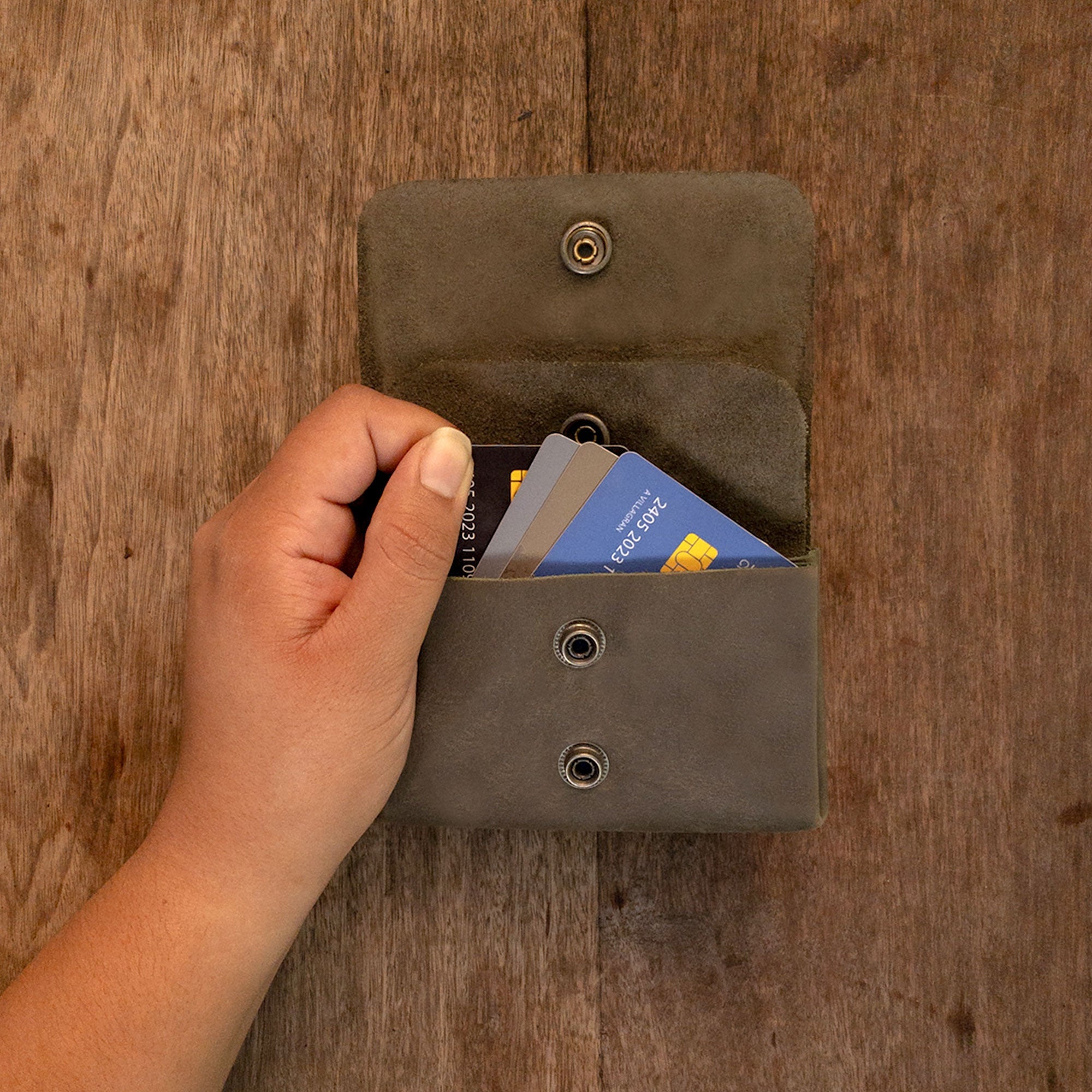 A hand using the olive Double Pouch Card Wallet by Hide & Drink to take out cards. This Leather Double Pouch Card Wallet is ideal as a card organizer, cash holder, and coin pouch, appealing to men and women, made from Full Grain Leather in La Antigua Guatemala & Oaxaca Mexico.