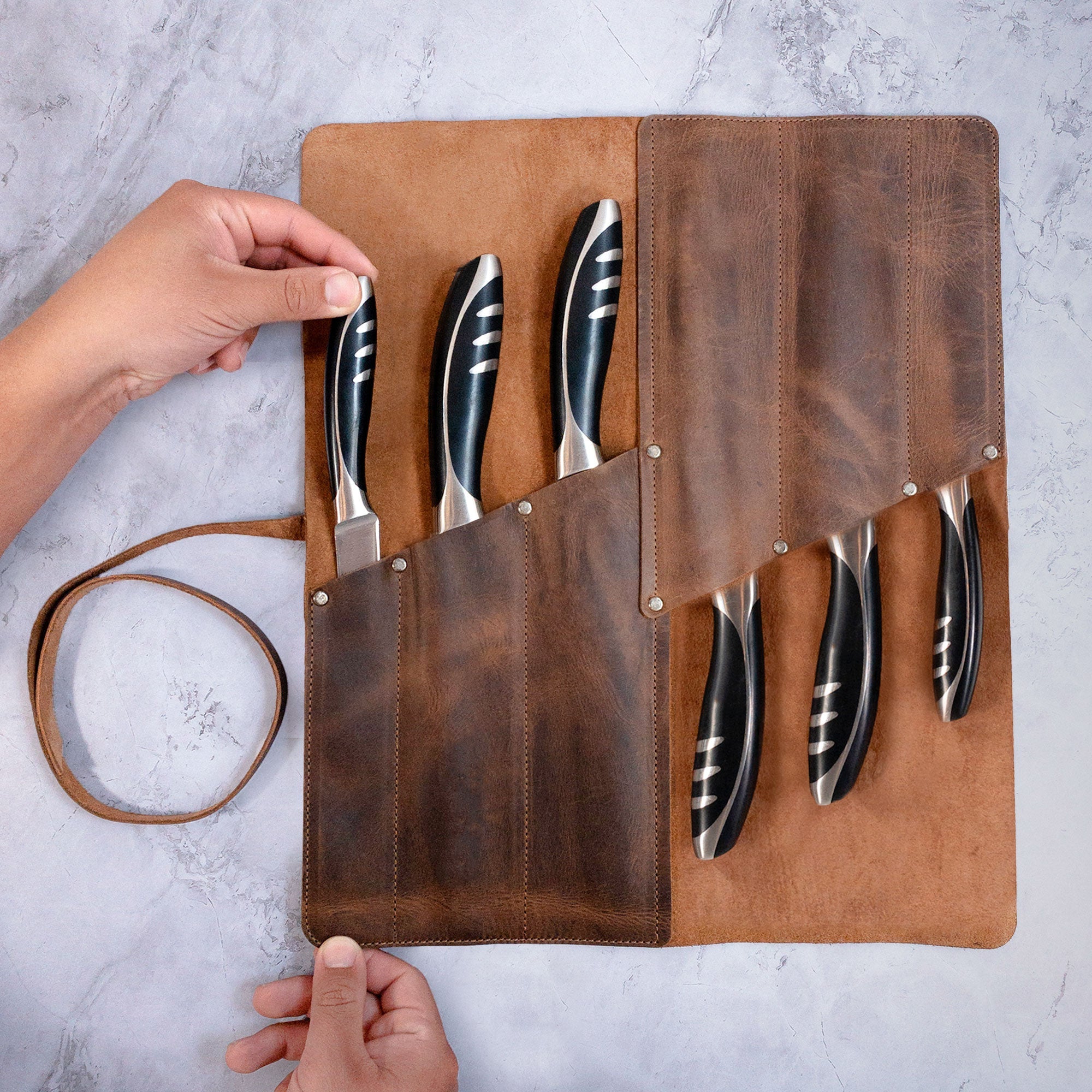 A user holding the open Cast Leather Co. Rectangular Knife Case with 6 Slots showcasing its interior with knives. This Portable Knife Organizer Case is designed for chefs on the go. Crafted from Full Grain Leather, it's an essential travel bag and cooking accessory. StockyardX delivers quality through small batch production with hand cut and hand sewn techniques, finished with a fire branded touch, and made in La Antigua Guatemala & Oaxaca Mexico.