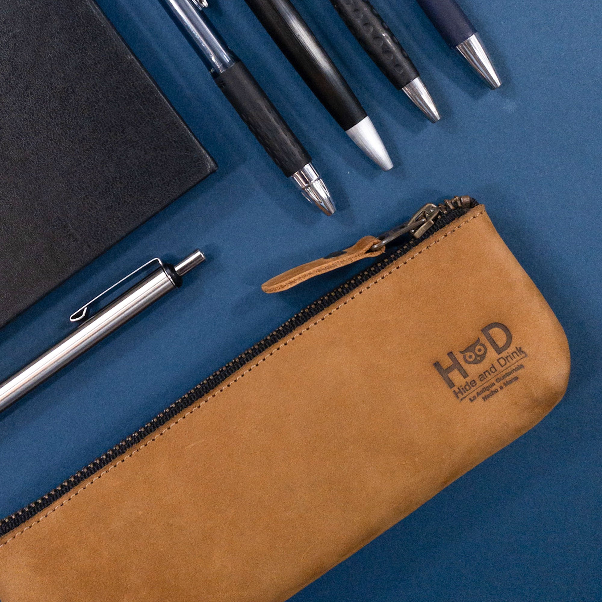 Pencil Pouch by Hide & Drink with various pens arranged nearby. This flat zippered holder is a rustic rectangular pencil case crafted from full grain leather, suitable for men and women. Handcrafted in La Antigua Guatemala, it is a classic style for artists.