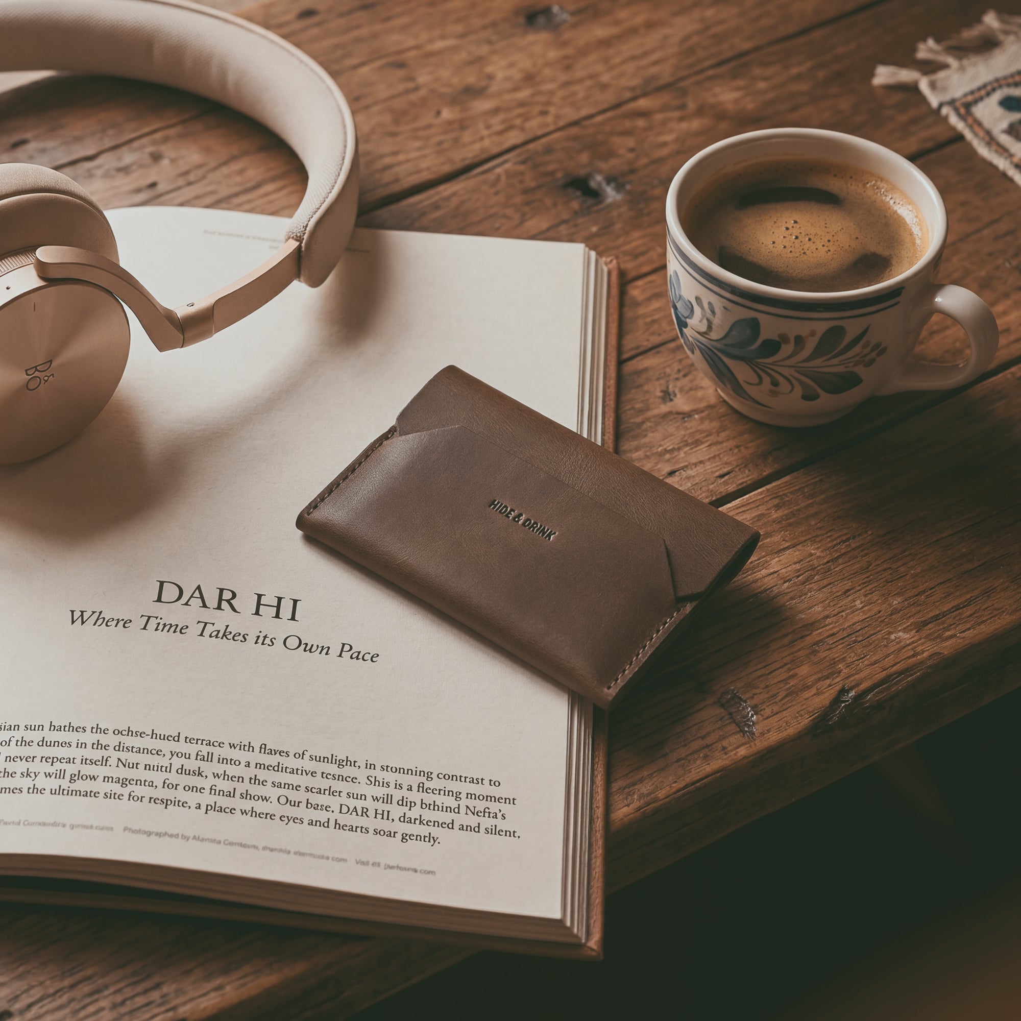 The Enfold Card Wallet by Hide & Drink styled amidst a cozy setting, showing its sleek design as a leather card wallet. Ideal for front pocket carrying, this compact credit card holder for men doubles as a minimalist thin business card organizer, with ample space for cash storage. Crafted from full grain leather, it exemplifies small batch production, hand cut and hand sewn, along with fire branding, showcasing the artisan touch from La Antigua Guatemala and Oaxaca Mexico.