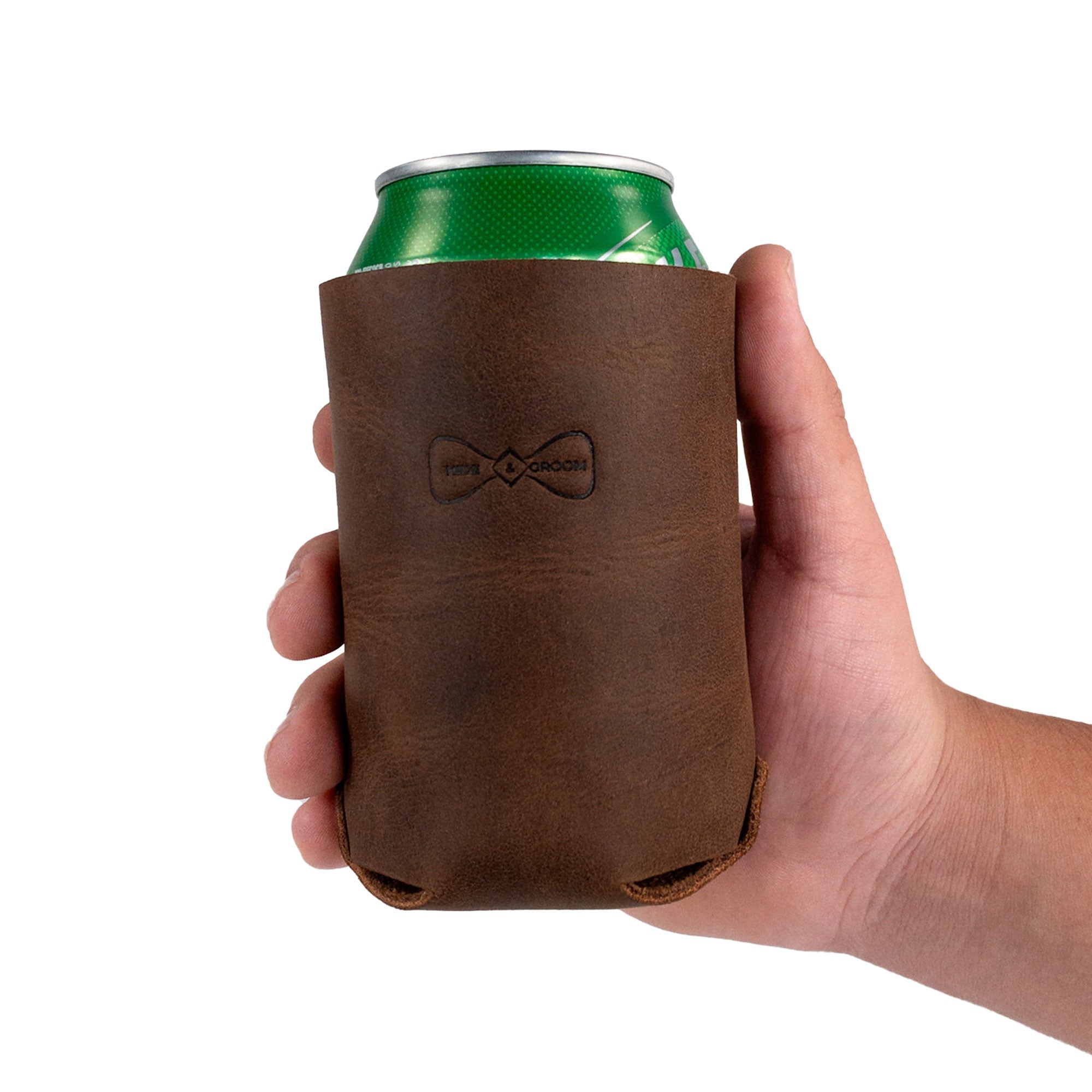 A hand holding the Hide & Groom Beer Can Sleeve (Set of 2), crafted from full grain leather. These collapsible rustic sleeves make ideal accessories for weddings and special events. Handmade in La Antigua Guatemala & Oaxaca Mexico, they feature hand cut and hand sewn techniques and are perfect for standard cans.