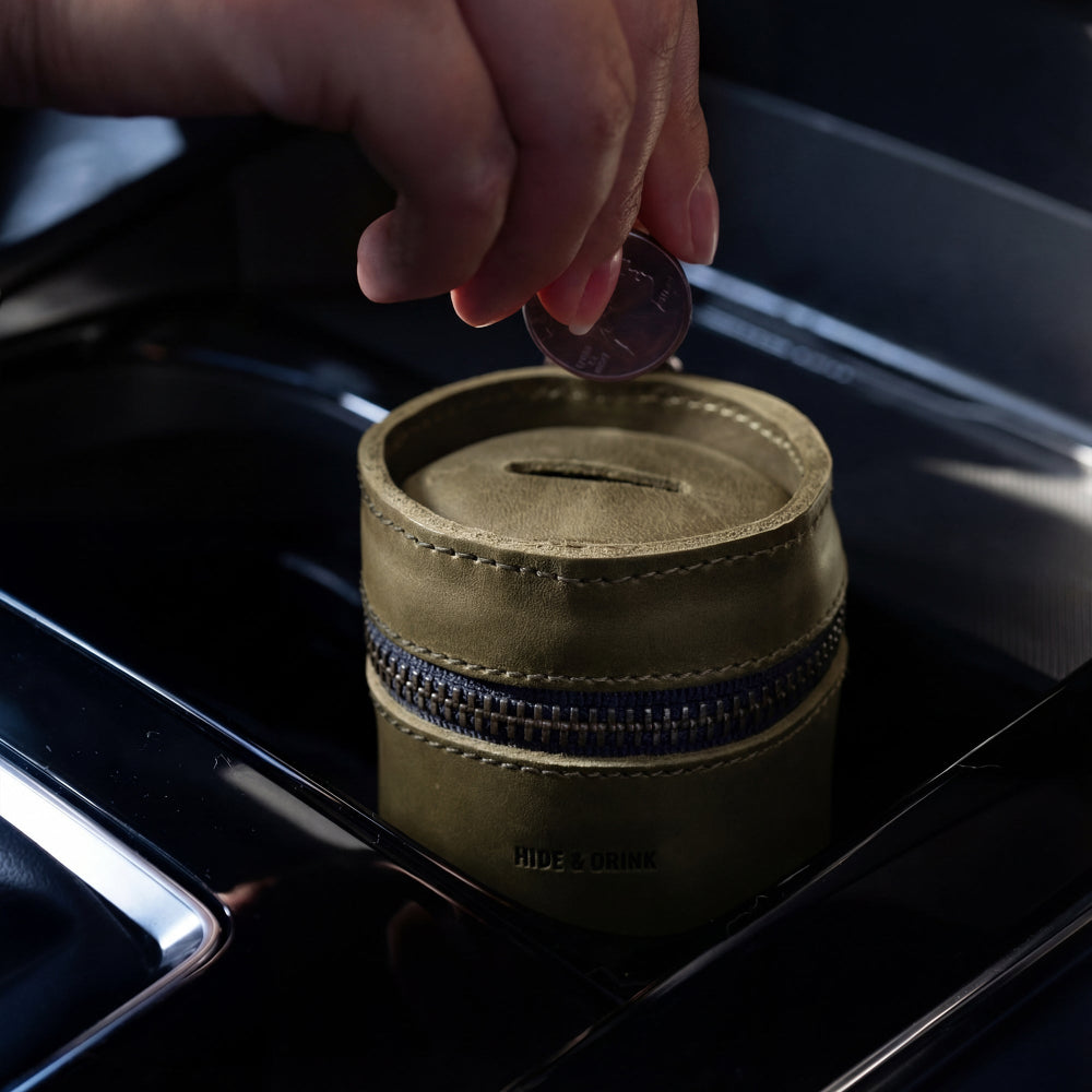 A hand inserting a coin into the Bucket Coin Release by Hide & Drink, demonstrating its practical use as a piggy bank bucket. This full grain leather cash organizer is a perfect small batch production item crafted by artisans in La Antigua Guatemala & Oaxaca Mexico.