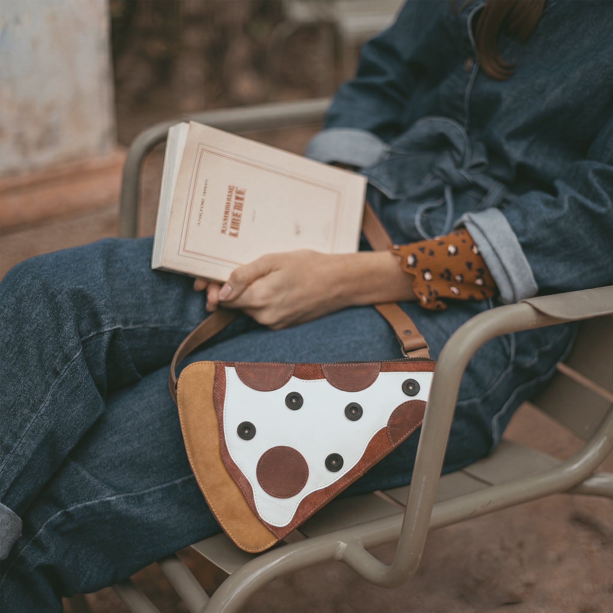 A model holding a Pizza Slice-Shaped Shoulder Bag from Tudi Frudi while enjoying a slice of pizza. The bag, designed in Full Grain Leather, highlights the artisan craftsmanship of small batch production and hand sewing, making it a perfect crossbody accessory.