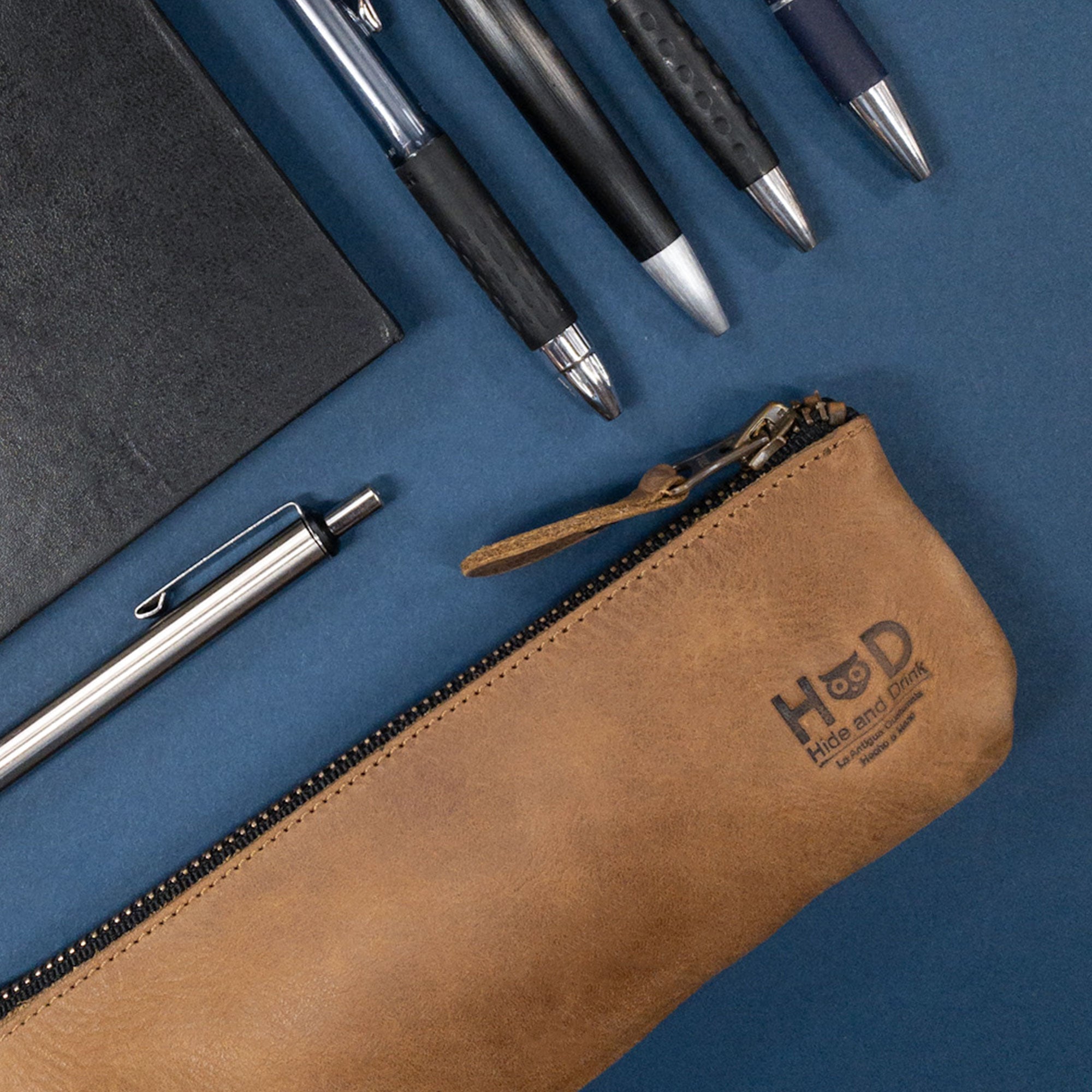 Pencil Pouch by Hide & Drink elegantly laid next to pens and a notebook. This rectangular pencil case, crafted from full grain leather, serves as a durable pen and marker pouch for men and women. Features a flat zippered holder, perfect for artists and handcrafted in La Antigua Guatemala.