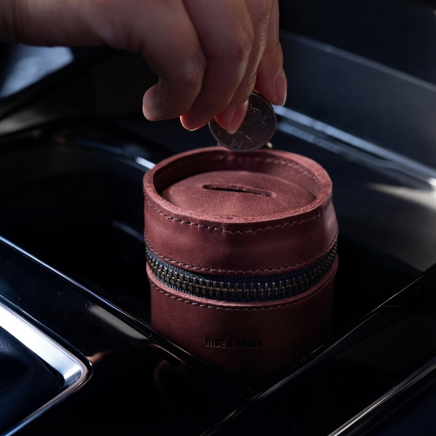 A person placing a coin into Hide & Drink's Bucket Coin Release, perfectly fitting within a car's console. This image showcases how the handy cash organizer, crafted from full grain leather, serves as an ideal money box and zippered pouch for everyday use.