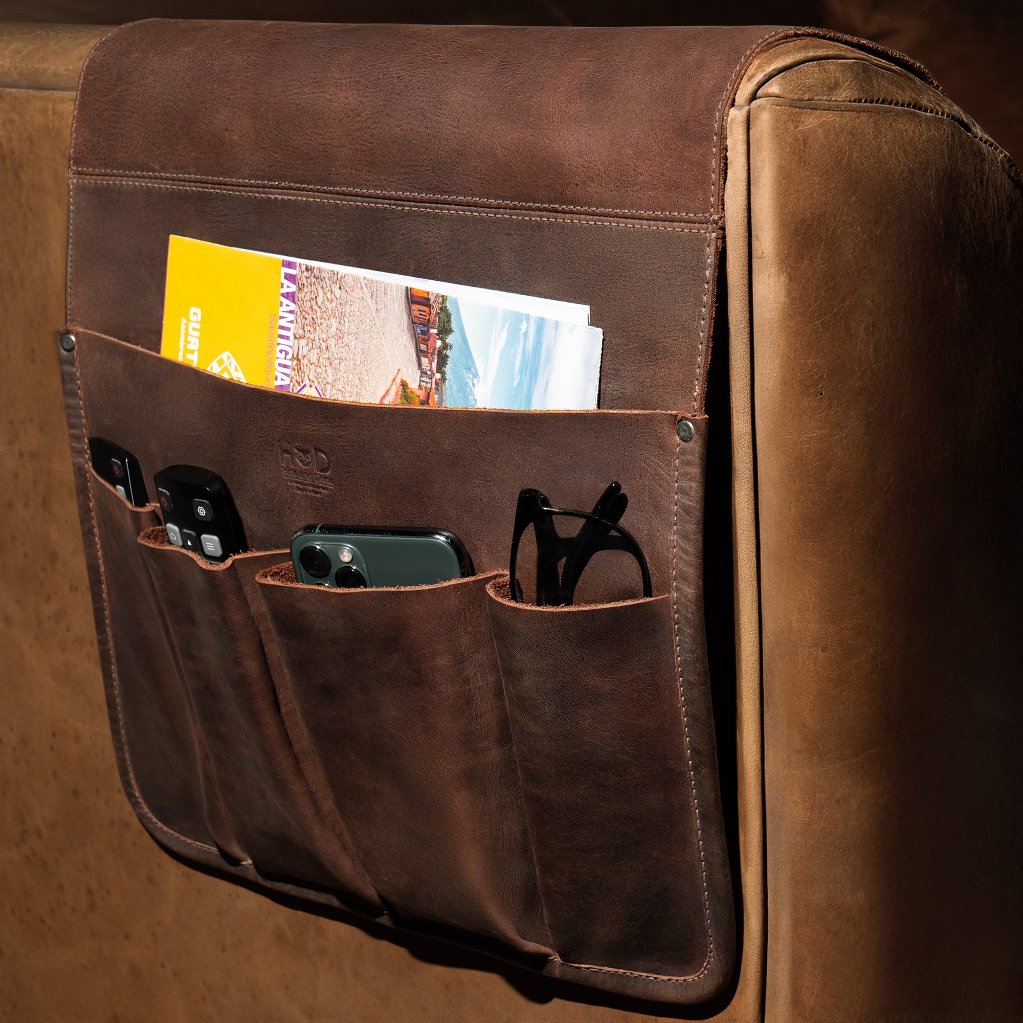 Hide & Drink's Remote Control Holder attached to a couch, elegantly displaying its practical design with pockets for remotes and magazines. A perfect addition to any living room, this armchair caddy is handmade, reflecting the small batch production quality.