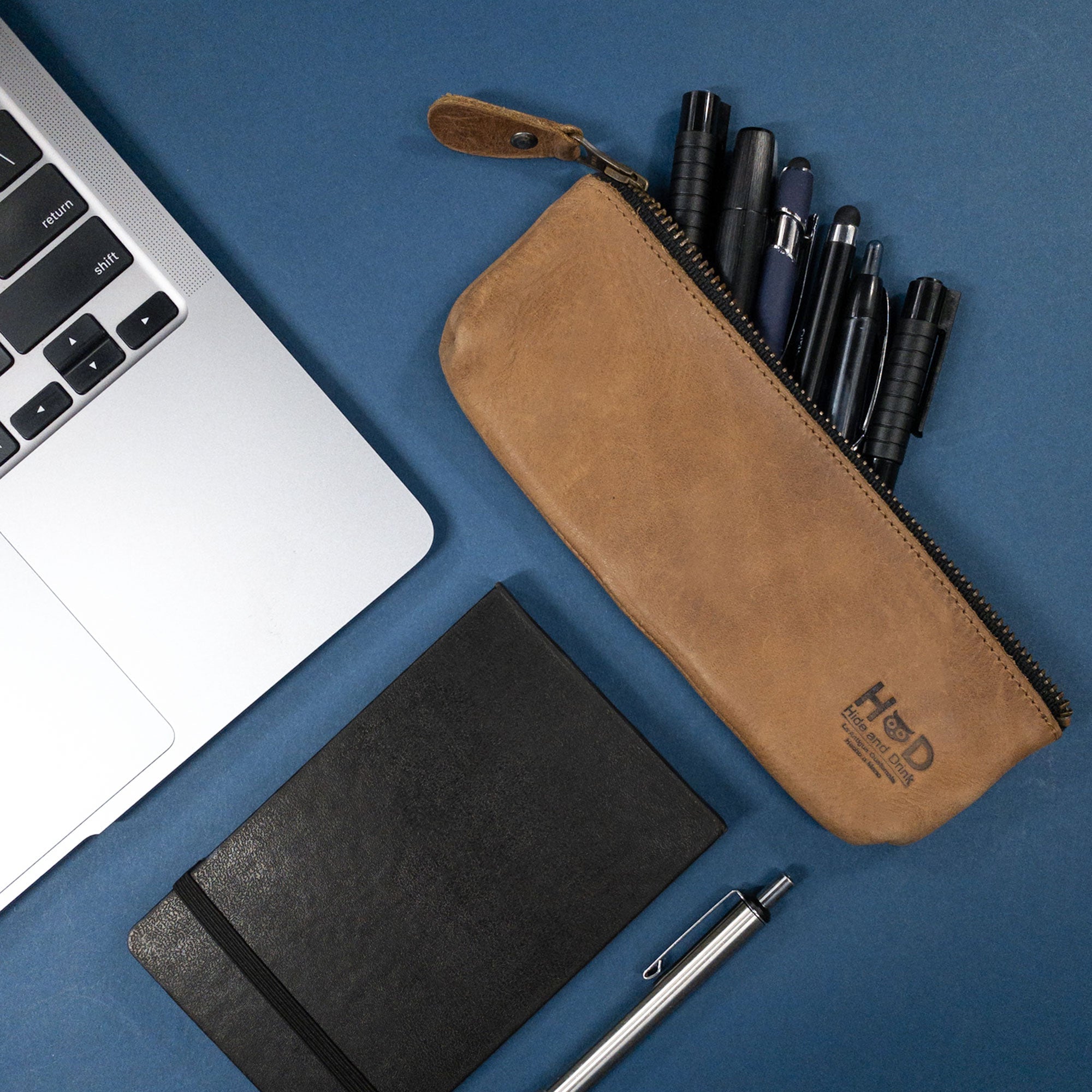 Stylish Pencil Pouch by Hide & Drink with pens inside, placed next to a laptop. This flat zippered holder is a rectangular pencil case made from full grain leather, designed for both men and women. A true classic for artists, handcrafted and fire branded by artisans in La Antigua Guatemala.