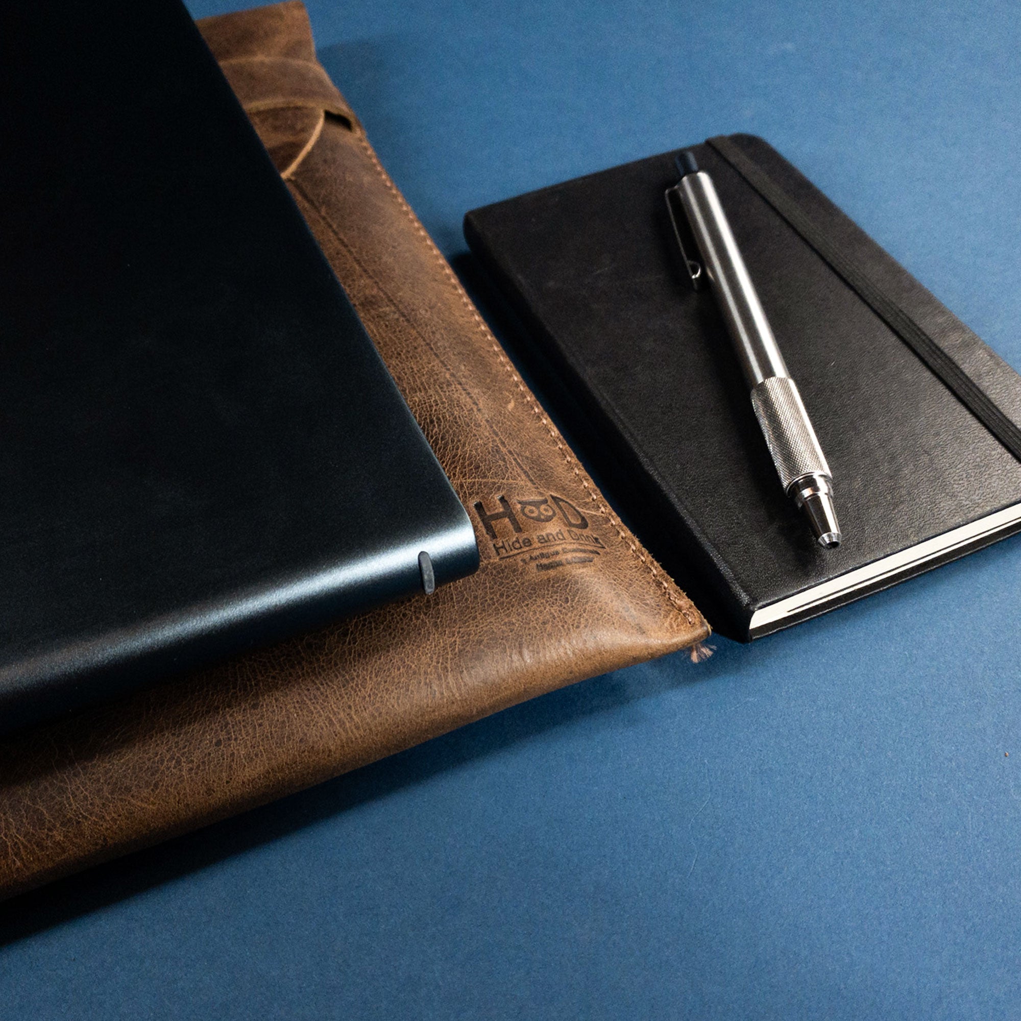 Lifestyle shot of the Hide & Drink MacBook Slim Case alongside a notebook and pen, perfect for the creative professional. This case is designed for MacBook Air/Pro 13 Inch with M1 Chip, crafted from full grain leather with artisan quality that showcases hand cut and hand sewn details.