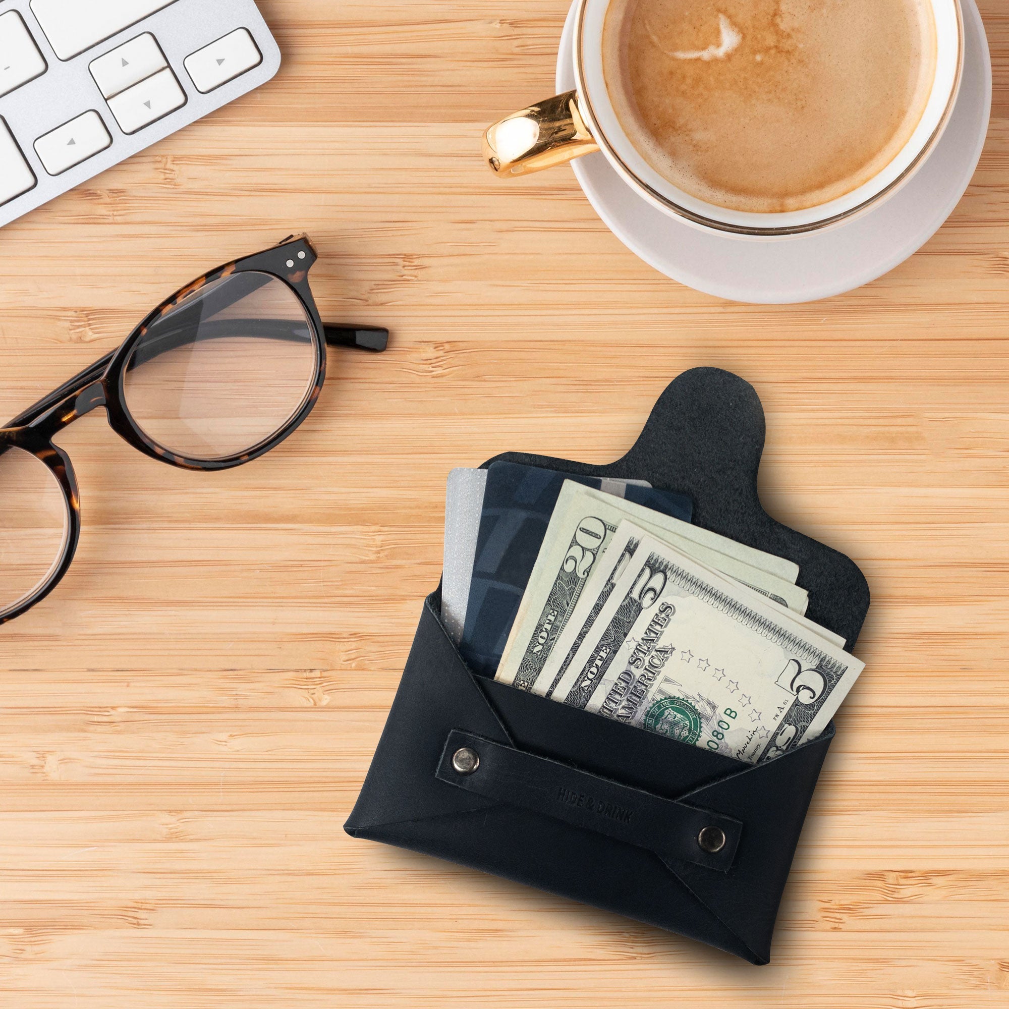 A Snapless Business Card Holder by Hide & Drink displayed alongside a coffee and glasses, emphasizing its versatility. This vintage wallet for men and women is designed to be a sturdy pouch and money organizer, crafted from luxurious Full Grain Leather, representing artisan craftsmanship from La Antigua Guatemala and Oaxaca Mexico.