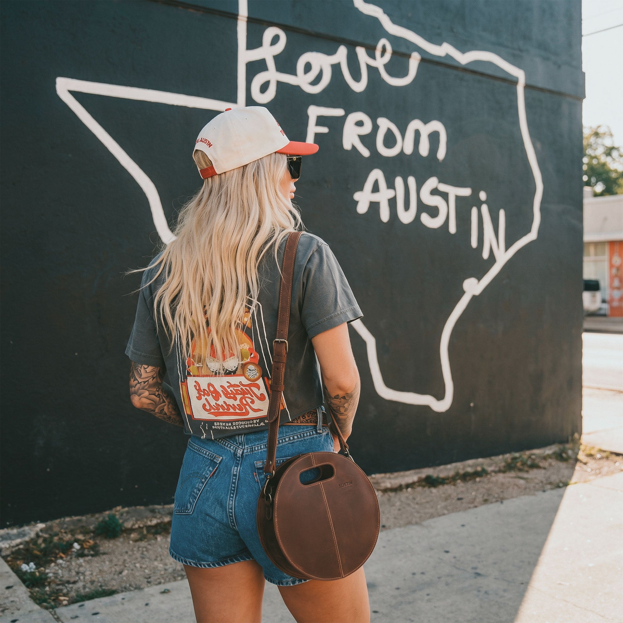 The Pecan Shoulder Bag by Austin Genuine, worn casually against a mural in Austin, Texas, exemplifying a unique blend of style and practicality, crafted from full grain leather with a focus on small batch production, hand cut and hand sewn, showcasing artisan work from La Antigua Guatemala & Oaxaca Mexico.