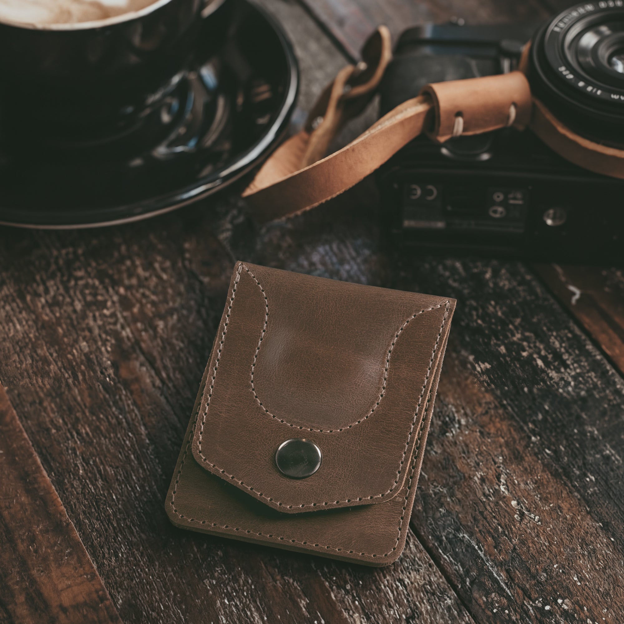The Card Holder with AirTag Slot by Hide & Drink rests on a table next to a cup of coffee, highlighting its full grain leather finish. This artisan-crafted vintage wallet is the ideal slim travel accessory for organizing your essentials with style.