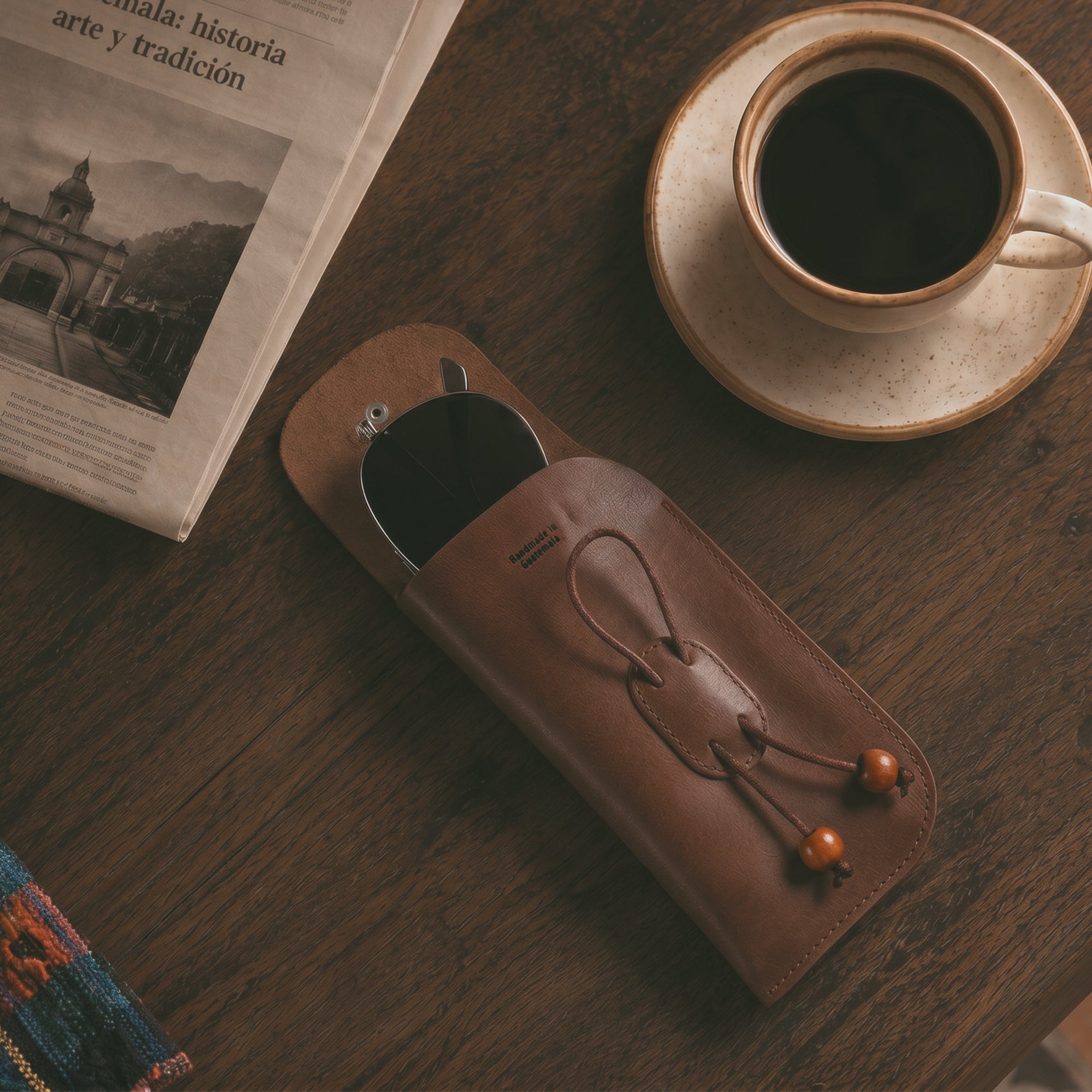 The Slim Eyeglasses Case by Hide & Drink, stylishly placed on a wooden table with a coffee cup. This leather glasses pouch is perfect for storing eyewear on the go, reflecting its high-quality full grain leather and artisan craftsmanship. A minimalistic eyewear cover, it highlights features from small batch production and is handmade in La Antigua Guatemala & Oaxaca Mexico.