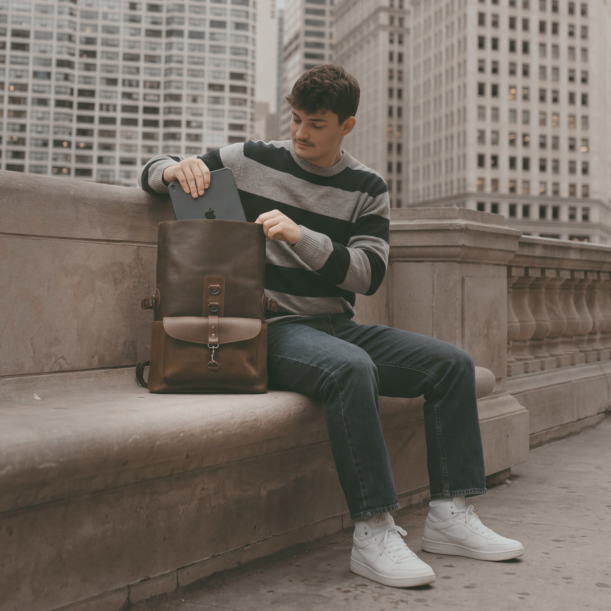 A person seated on a city bench with the Vintage Adventure Crossbody Bag from Littlefield Chicago. This messenger bag, ideal for urban adventures, showcases its practical design, handcrafted from premium materials in La Antigua Guatemala, making it a perfect travel sling bag.