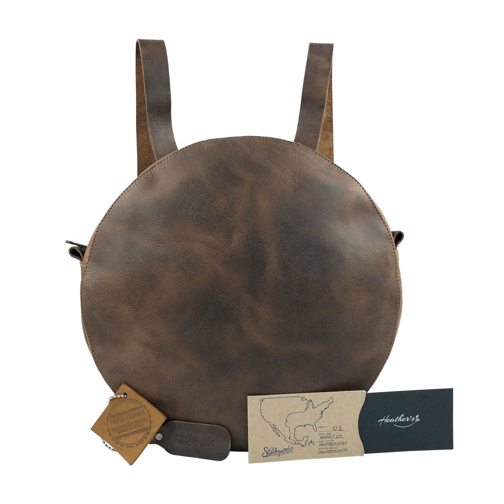 Heather's Circular Backpack presented with branding tags and artisan labels, showcasing its unique full grain leather craftsmanship. This stylish travel accessory functions beautifully as a casual daypack, integrating adjustable straps and emphasizing the handmade quality from artisans in La Antigua Guatemala & Oaxaca Mexico.